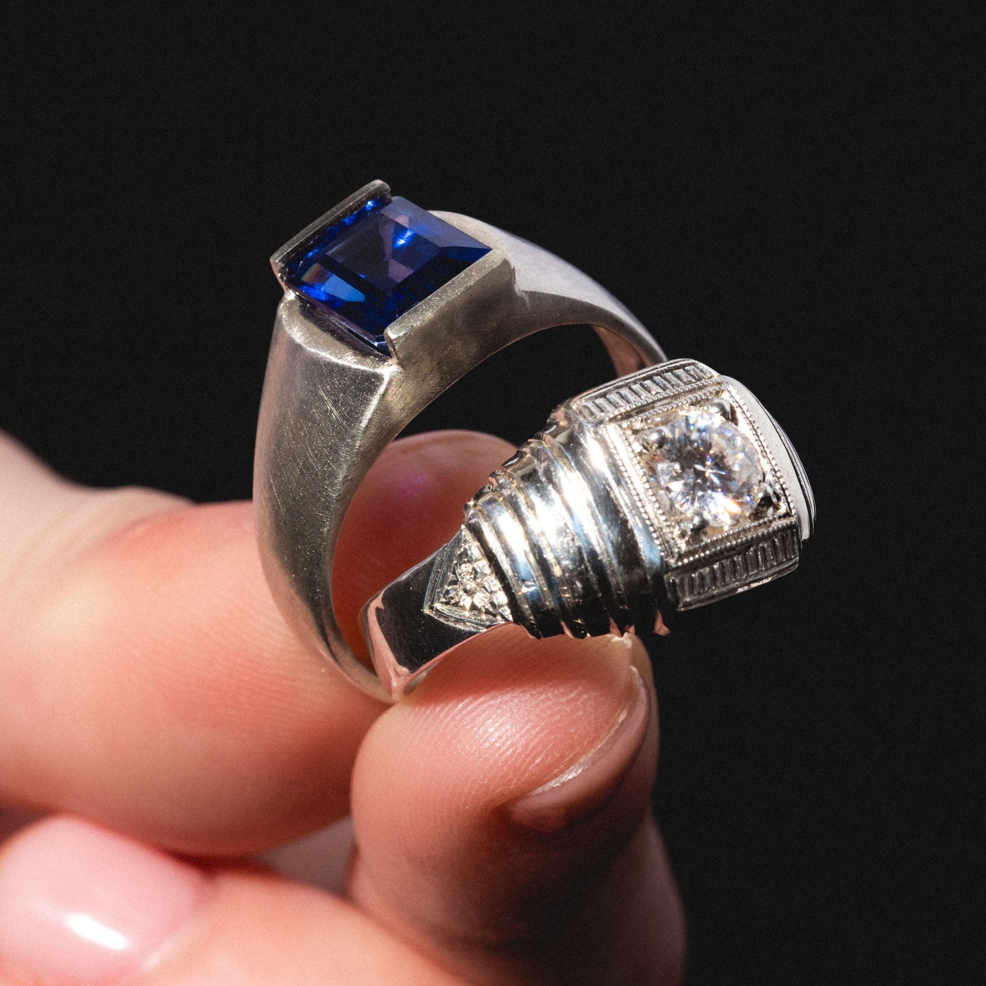 A hand displays the Sacred Ring, featuring a rectangular blue gemstone set in silver with a round clear stone in a decorative setting, against a black background.