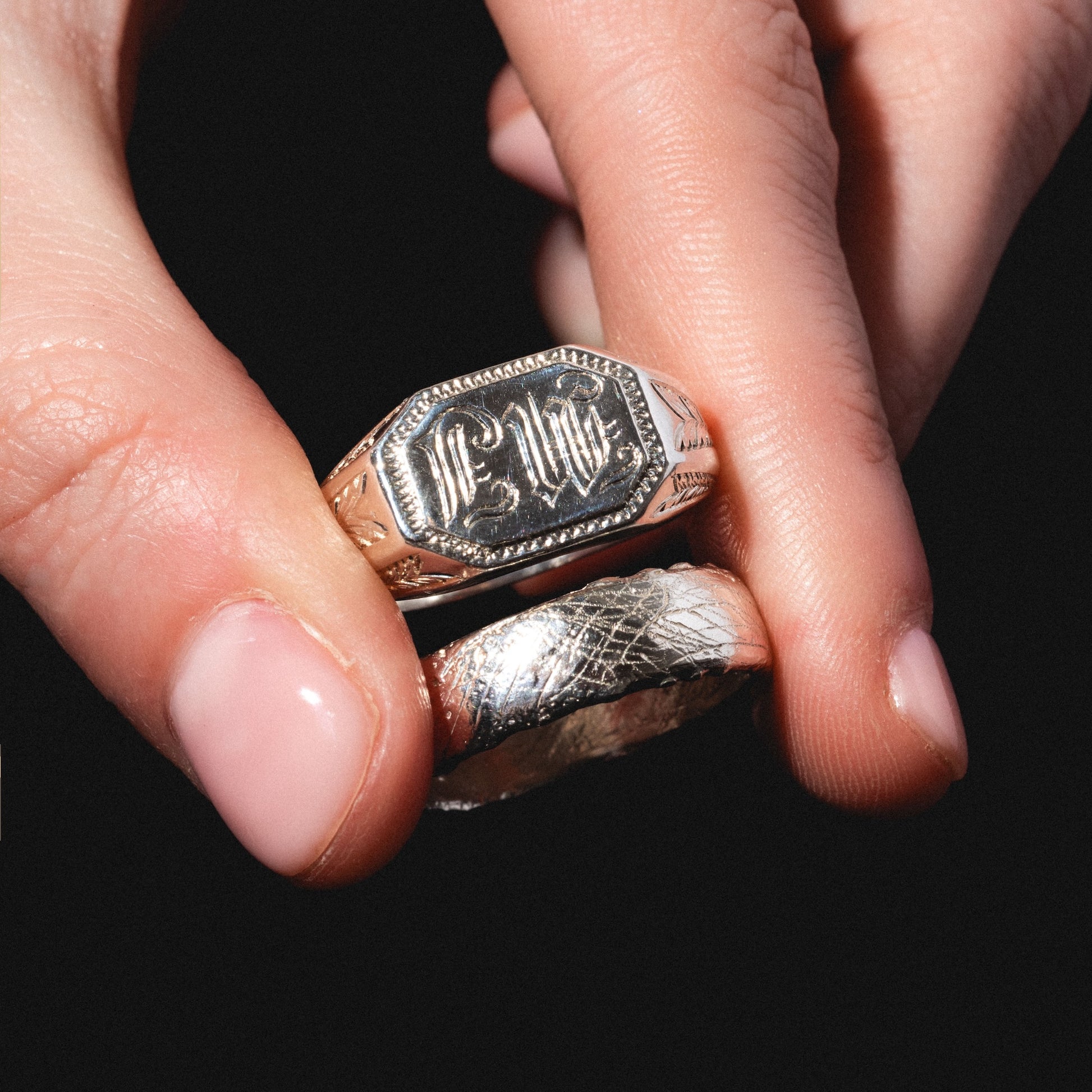 A close-up of a hand holding two silver rings against a black background: the Sovereign Signet Ring with an engraved design on its flat surface, and another featuring a worn, scratched texture.