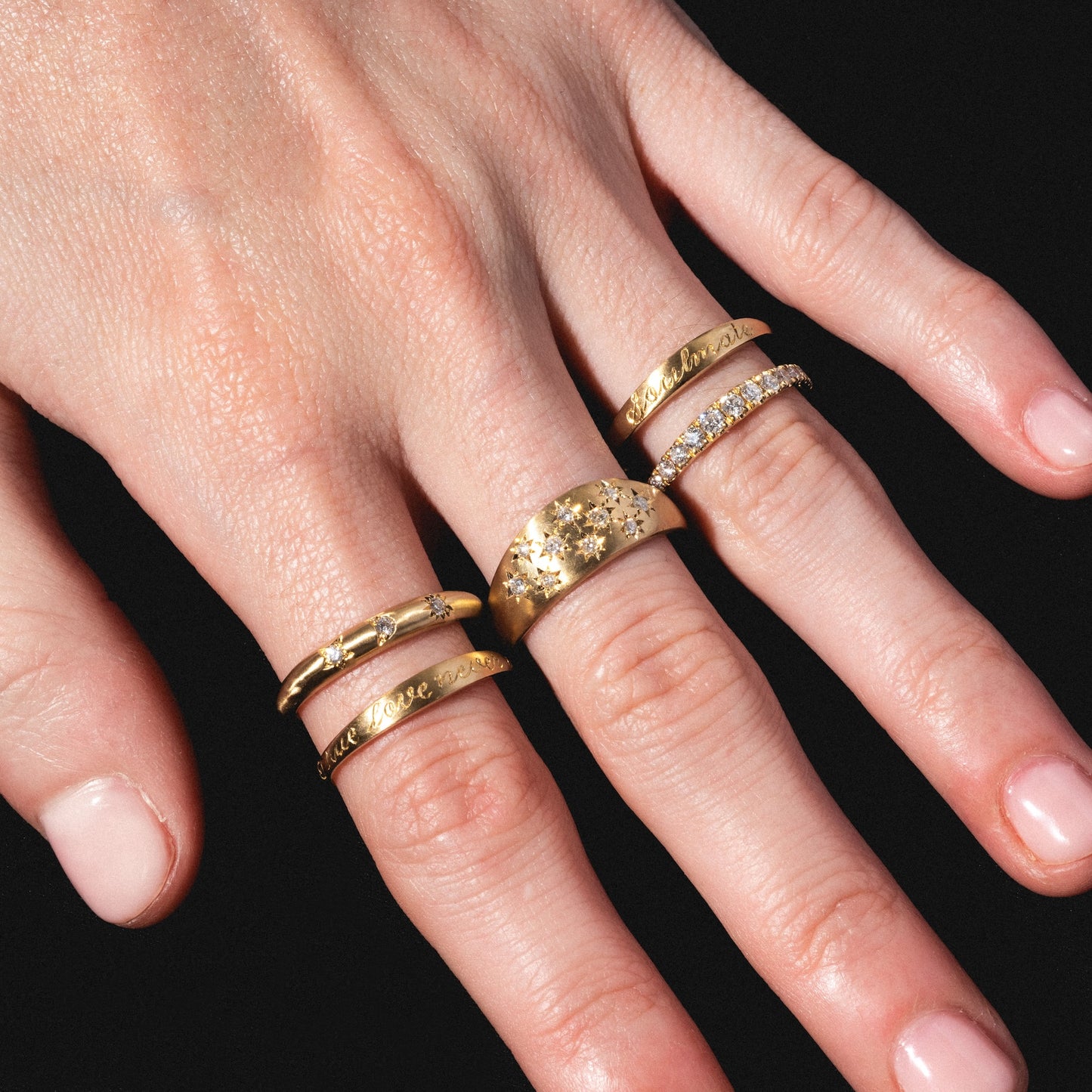 A close-up of a hand with light pink nail polish wearing the Soulmate Band, along with gold rings featuring engravings, star-shaped designs, and clear stones, set against a black background.