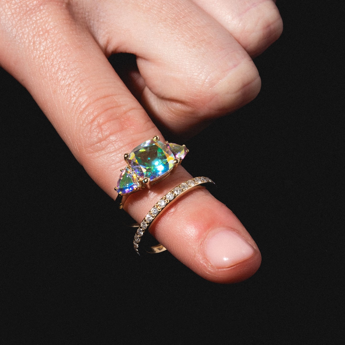 A hand with light pink nail polish wears the Seraphina Ring—gold, featuring a large iridescent gemstone flanked by two side stones—and a thin diamond band, shown against a black background.