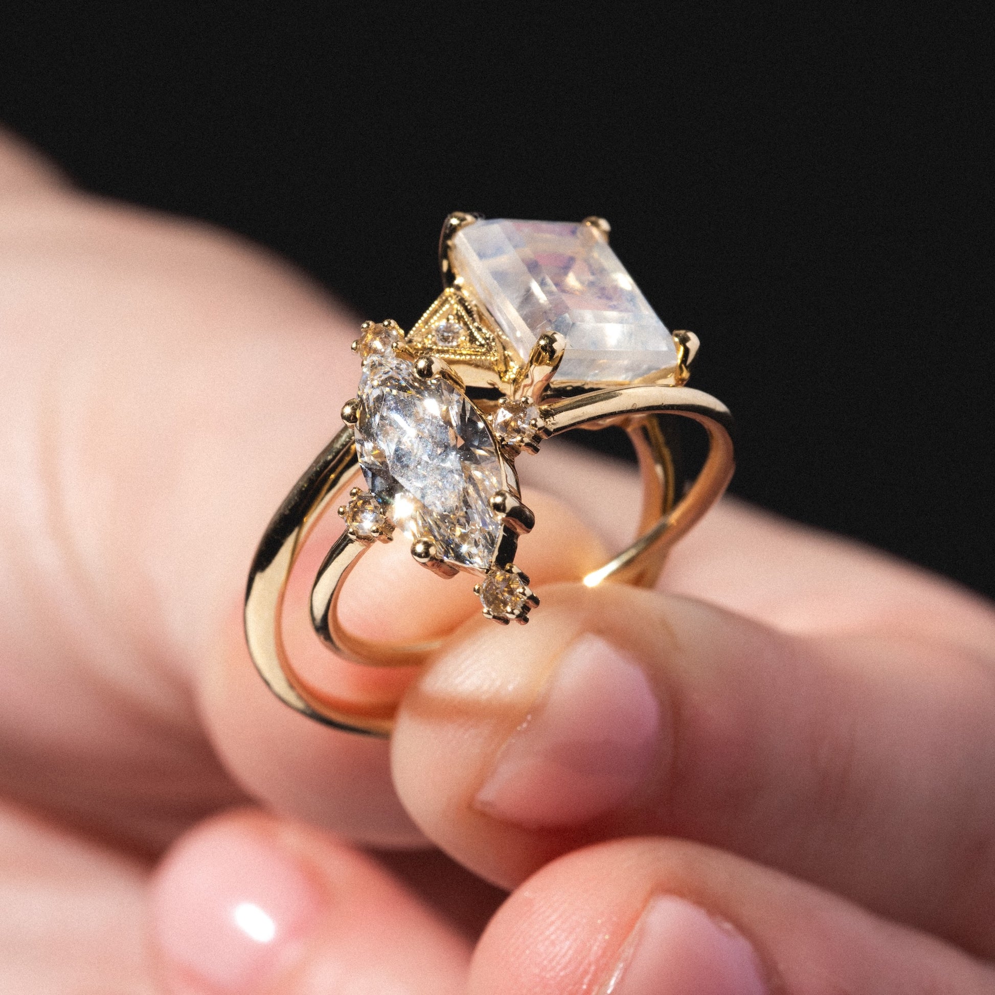 A hand holds two gold Celestia Rings—one with a large rectangular gemstone, the other with a marquise-cut stone and small round accents—set against a dark background.
