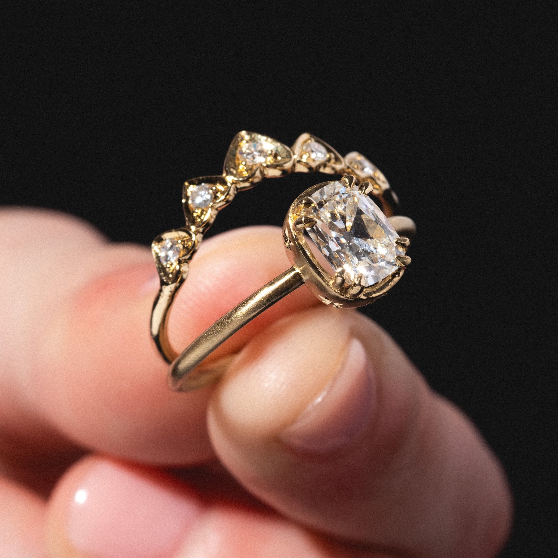 A hand holds two gold rings: one has a large oval diamond, and the other is the Radiant Hearts Band, set with five pear-shaped diamonds in a curved row. The background is black.