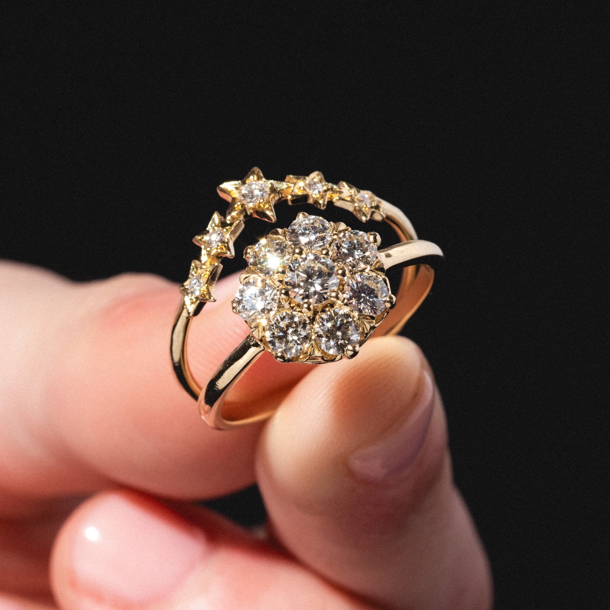 A hand displays the Stars Aligned Band: a gold ring set against a black background, featuring a large central cluster of round diamonds and a curved band adorned with small star-shaped accents.