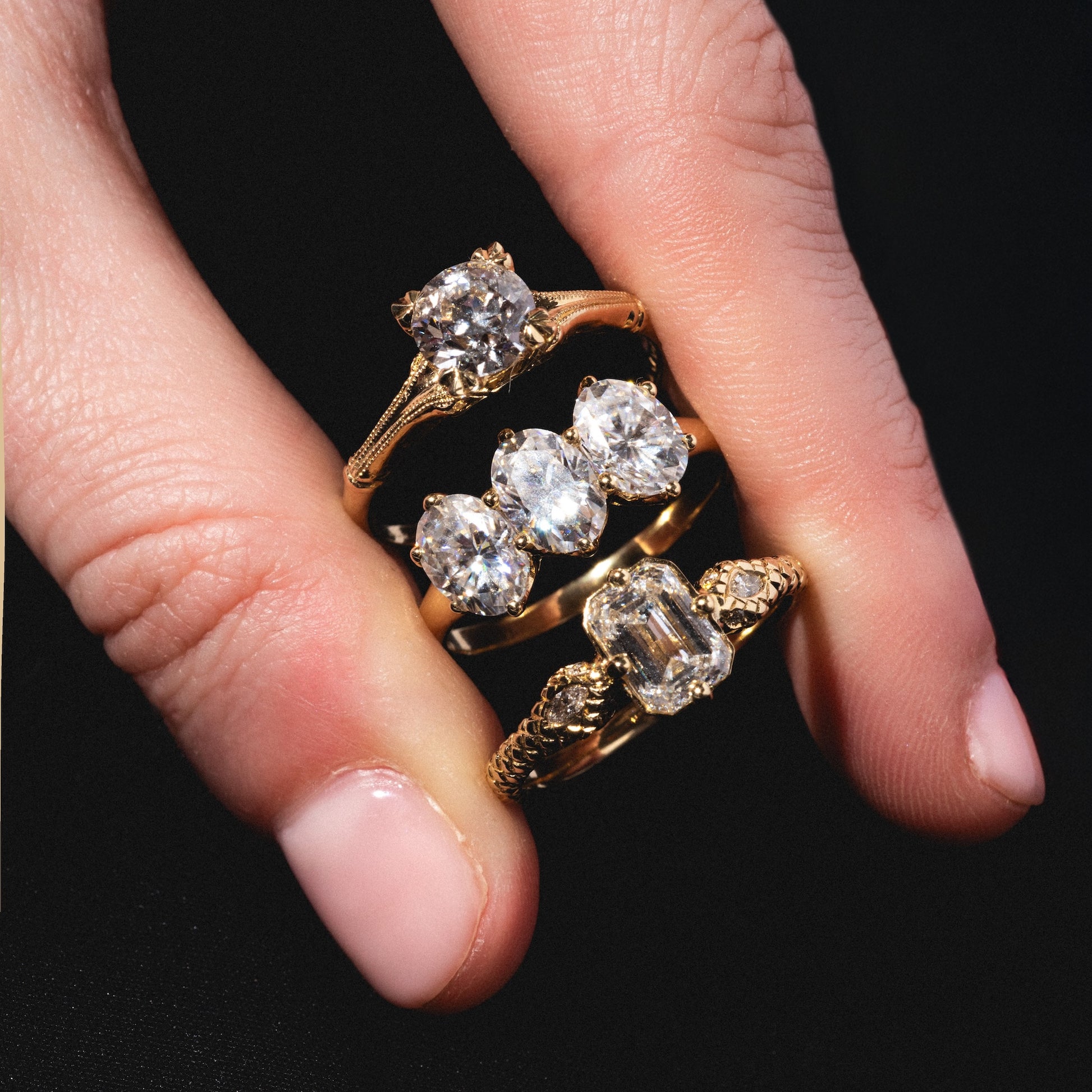 A hand with light pink nails displays three Enchantress Rings, each adorned with sparkling clear gemstones in unique settings, against a black background.
