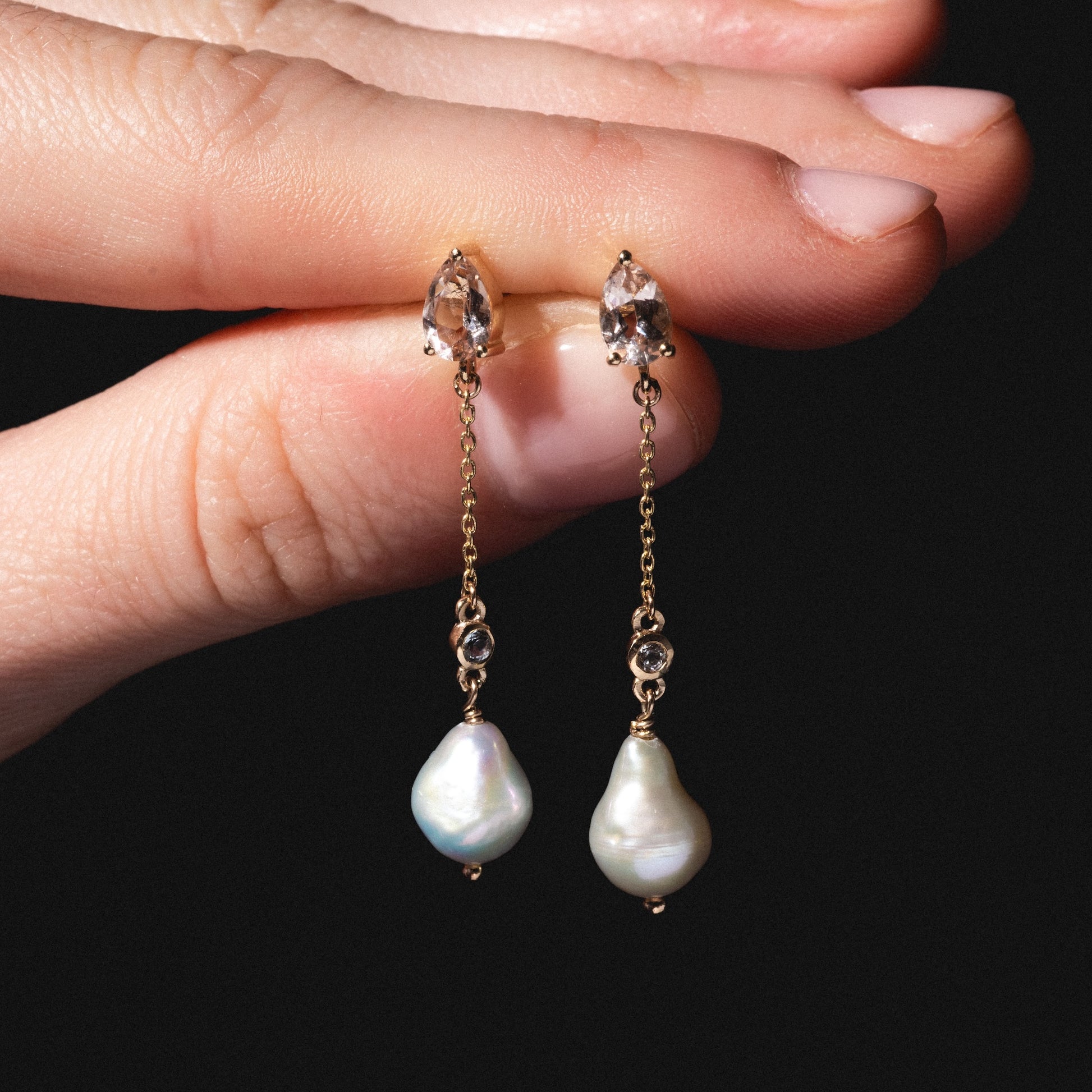 A hand holds Solid Gold Citrine and Pearl Droplet Earrings featuring teardrop citrine crystals, delicate chains, round stones, and irregular white pearls against a black background.