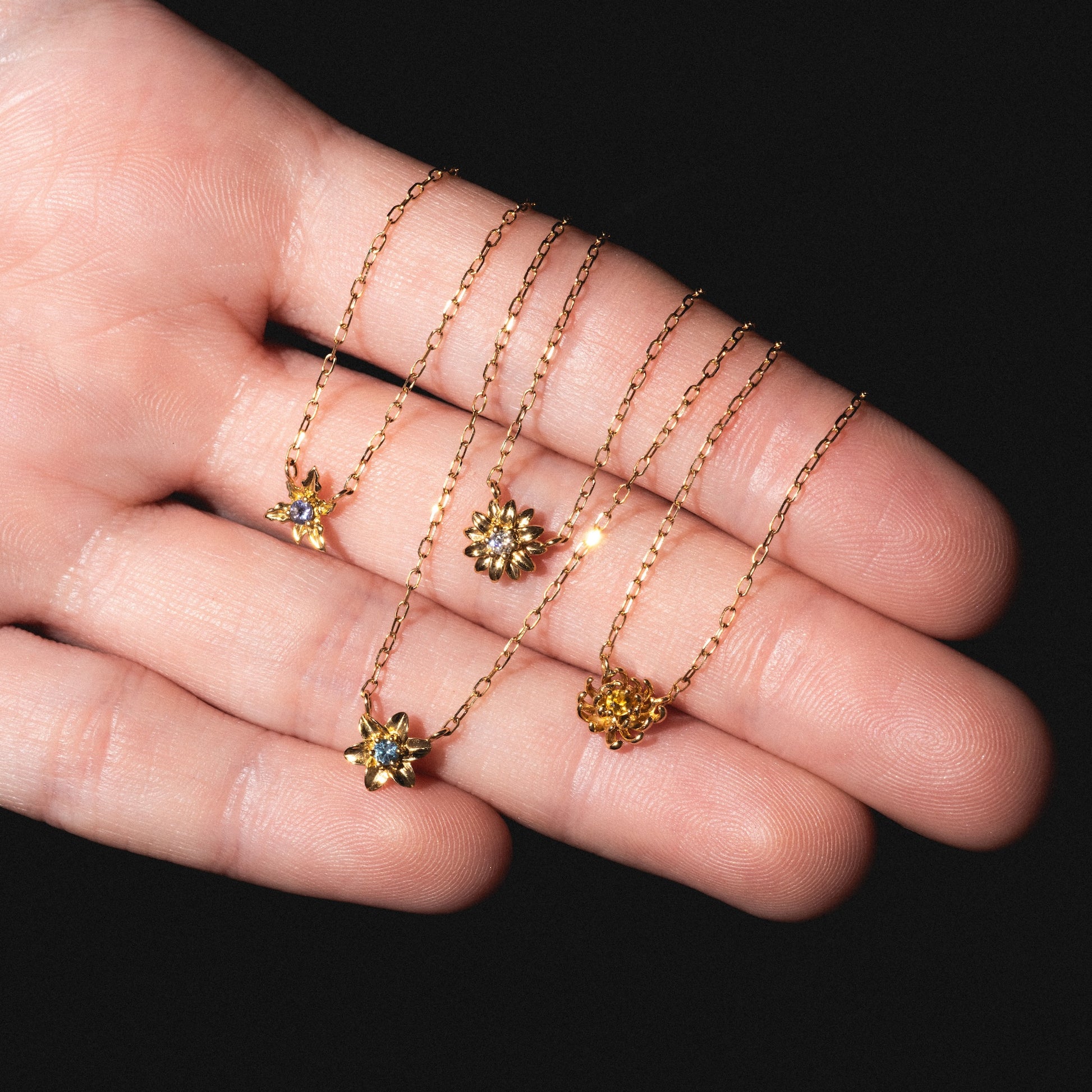 A hand displays four Solid Gold Birth Flower Bloom Necklaces, each featuring a unique, small flower-shaped pendant, against a black background.