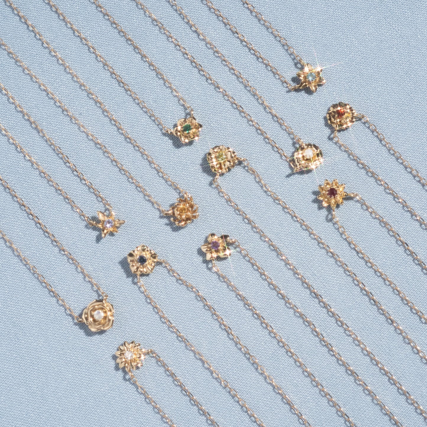 The Solid Gold Birth Flower Bloom Necklace, featuring an intricate floral pendant, is displayed neatly in parallel rows on light blue textured fabric.