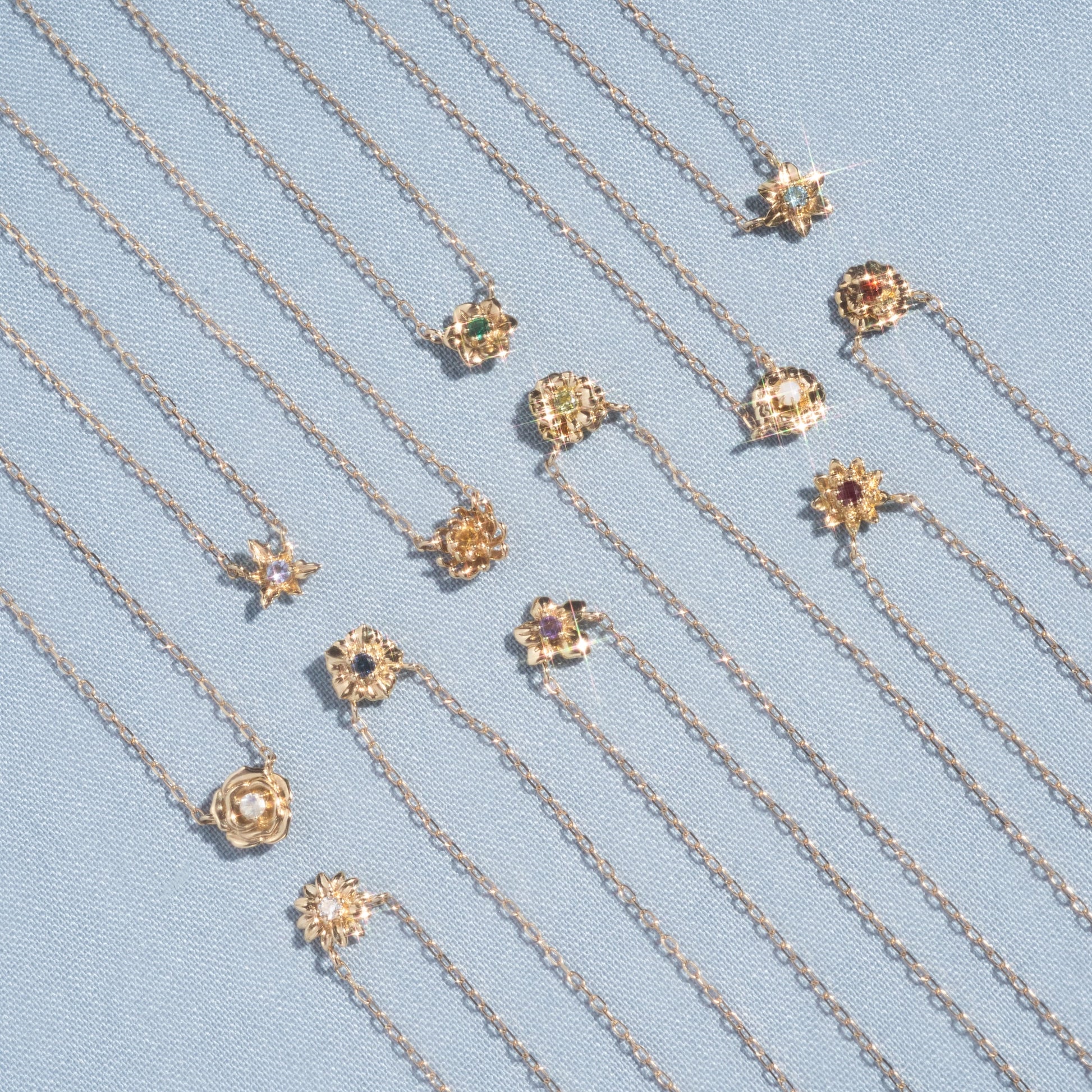 The Solid Gold Birth Flower Bloom Necklace, featuring an intricate floral pendant, is displayed neatly in parallel rows on light blue textured fabric.