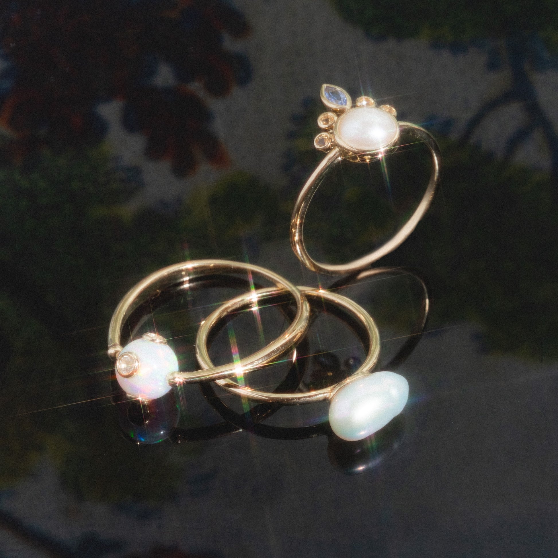 The Solid Gold Organic Pearl Solitaire Ring features iridescent gemstones and pearl-like accents, displayed on a dark, reflective surface with soft, colorful reflections and blurred floral patterns in the background.