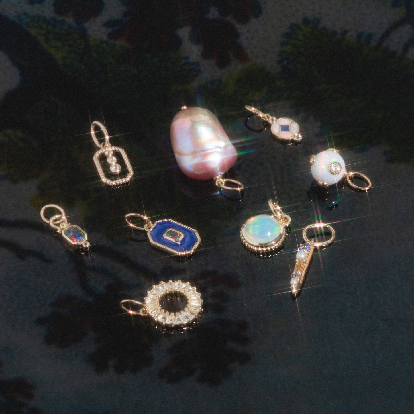 Solid Gold Statement Charm: A selection of small, sparkling gemstone and pearl charms in gold settings, including unique gold necklace charms, all arranged on a glossy dark floral background with glimmering reflections.
