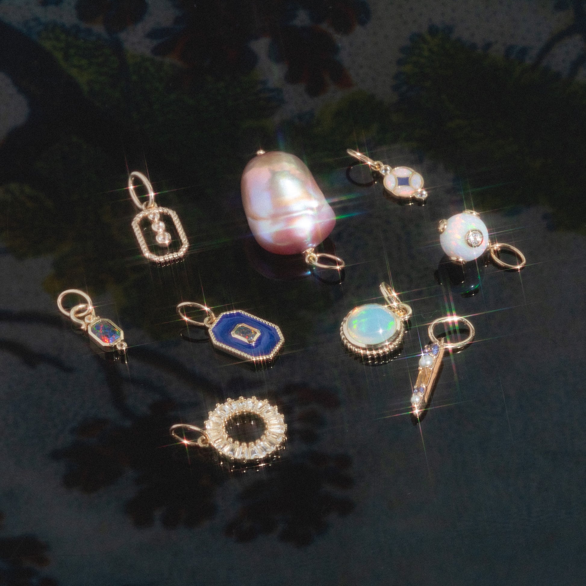 Solid Gold Statement Charm: A selection of small, sparkling gemstone and pearl charms in gold settings, including unique gold necklace charms, all arranged on a glossy dark floral background with glimmering reflections.