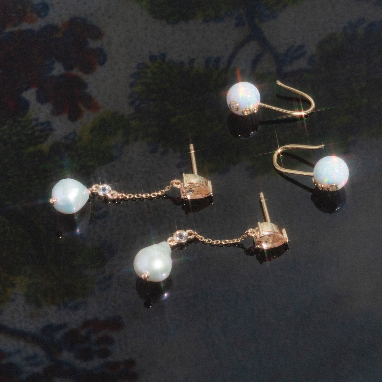 A pair of Solid Gold Citrine and Pearl Droplet Earrings displayed on a reflective dark surface with a blurred floral background.