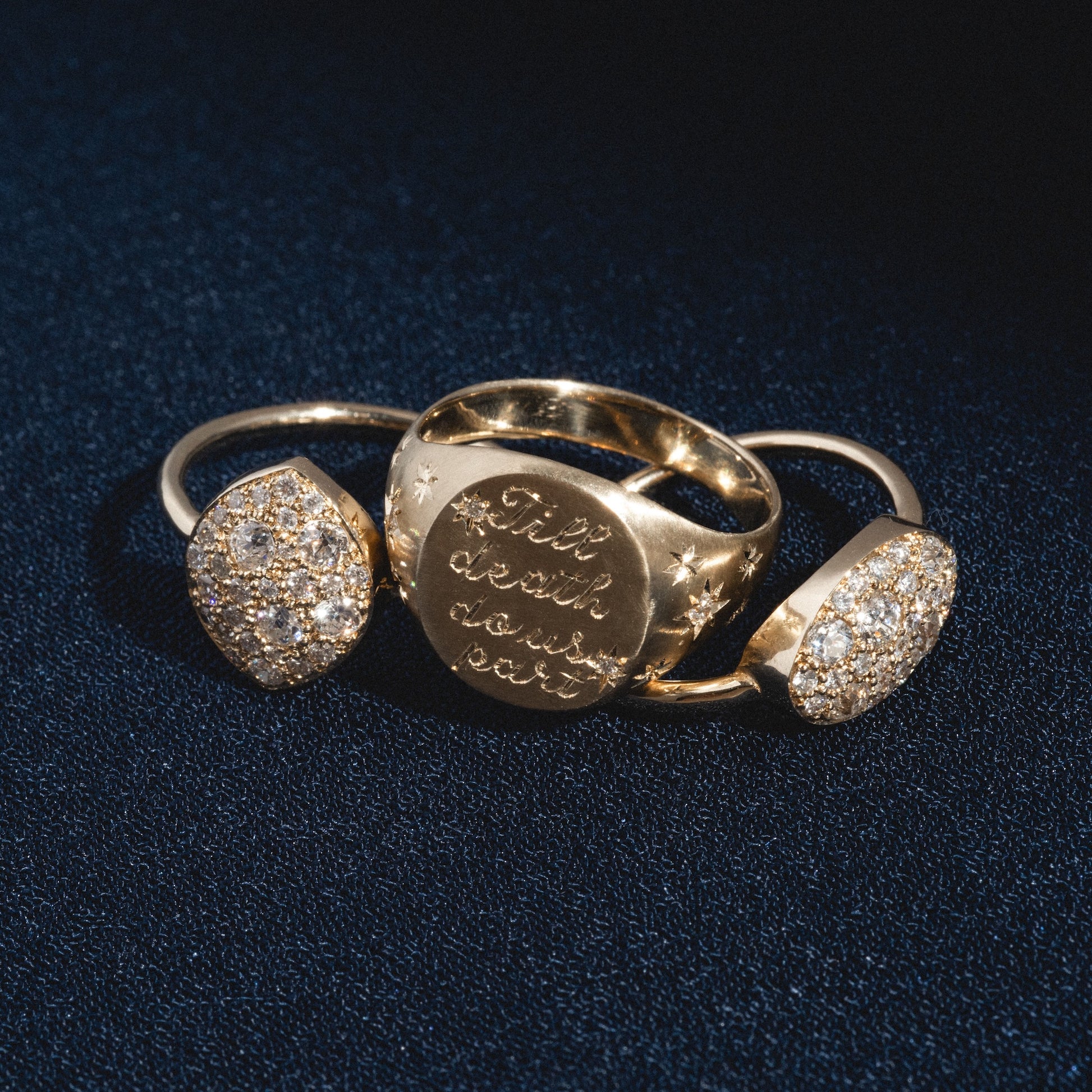 Three gold rings on a dark textured surface; the center is the Till Death Do Us Part Signet with Diamonds, engraved with "Til death does part" and star designs, flanked by two rings featuring clusters of sparkling stones.