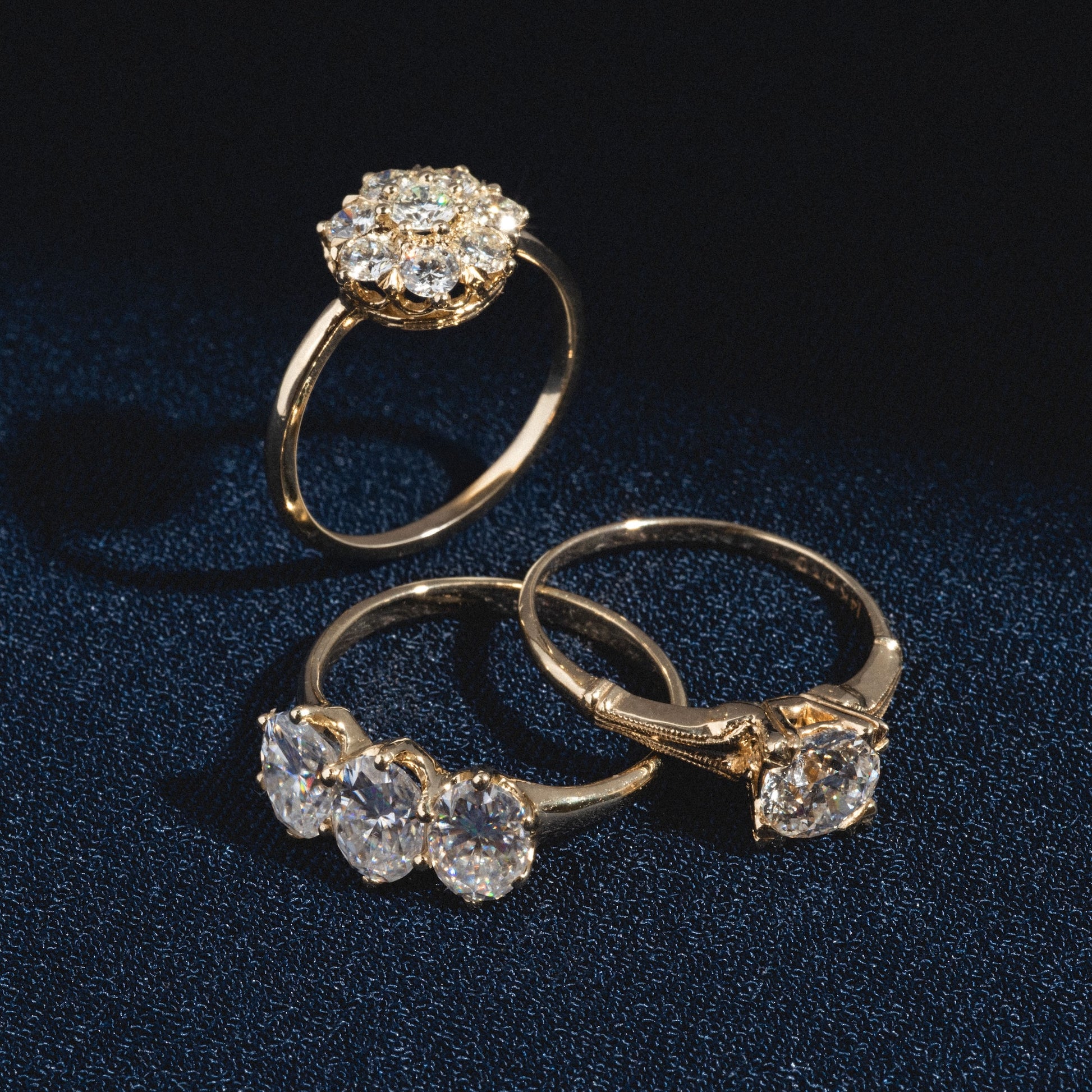 Three Queenie Rings with large, sparkling diamonds are displayed on dark blue fabric; two lie flat and one stands upright, all catching the light.
