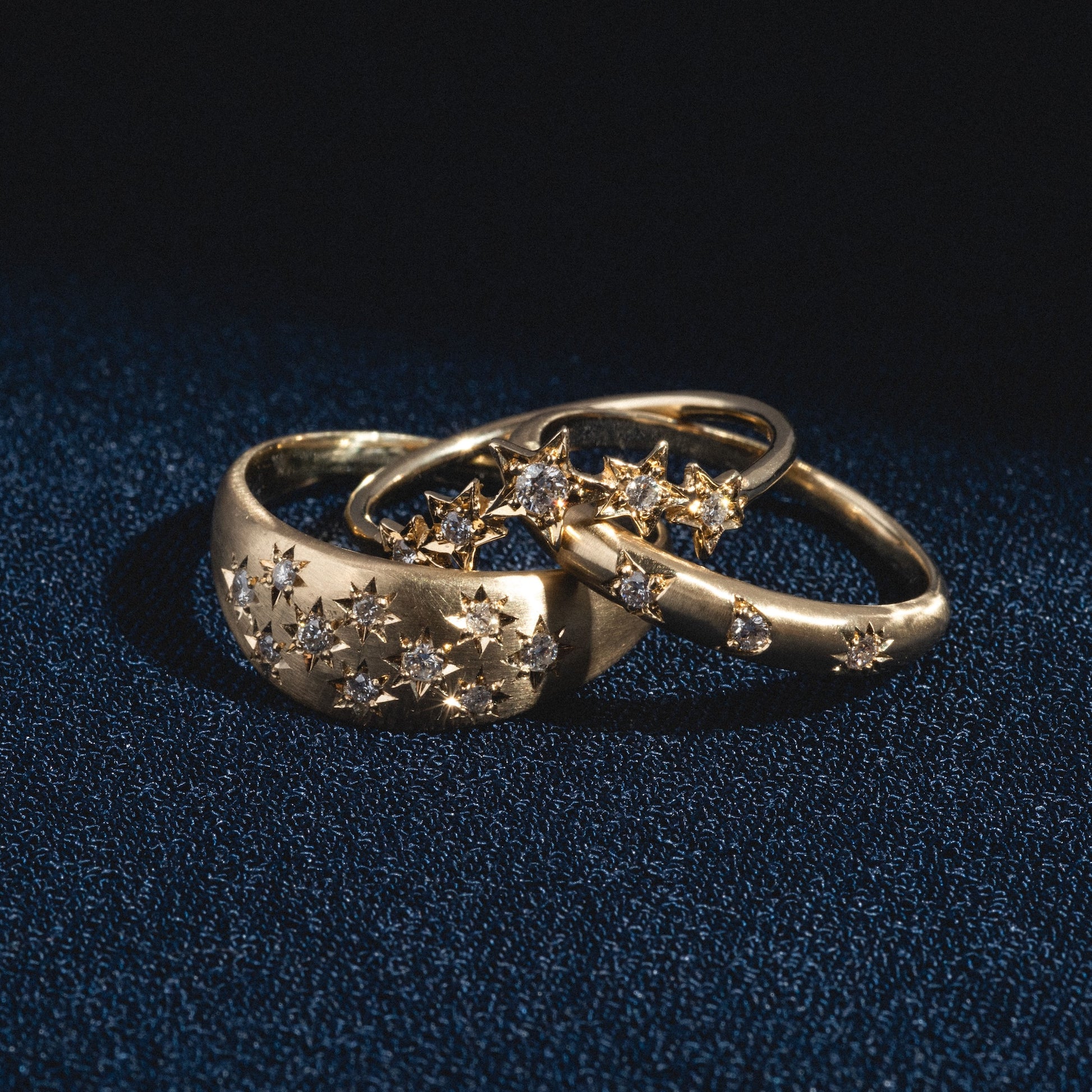 Three "You are my Sun, Moon and Star Band" gold rings with small star-shaped diamond clusters rest on a dark blue textured surface, illuminated by soft, focused lighting.
