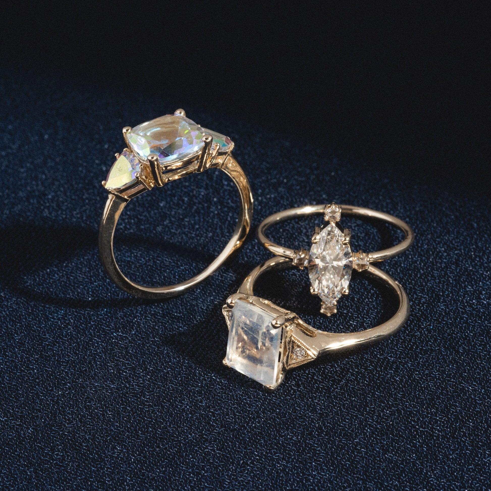 The Seraphina Ring, featuring large gemstones in oval, rectangular, and kite shapes, is displayed on a dark textured surface. Each stone catches the light, revealing a subtle iridescent sheen.