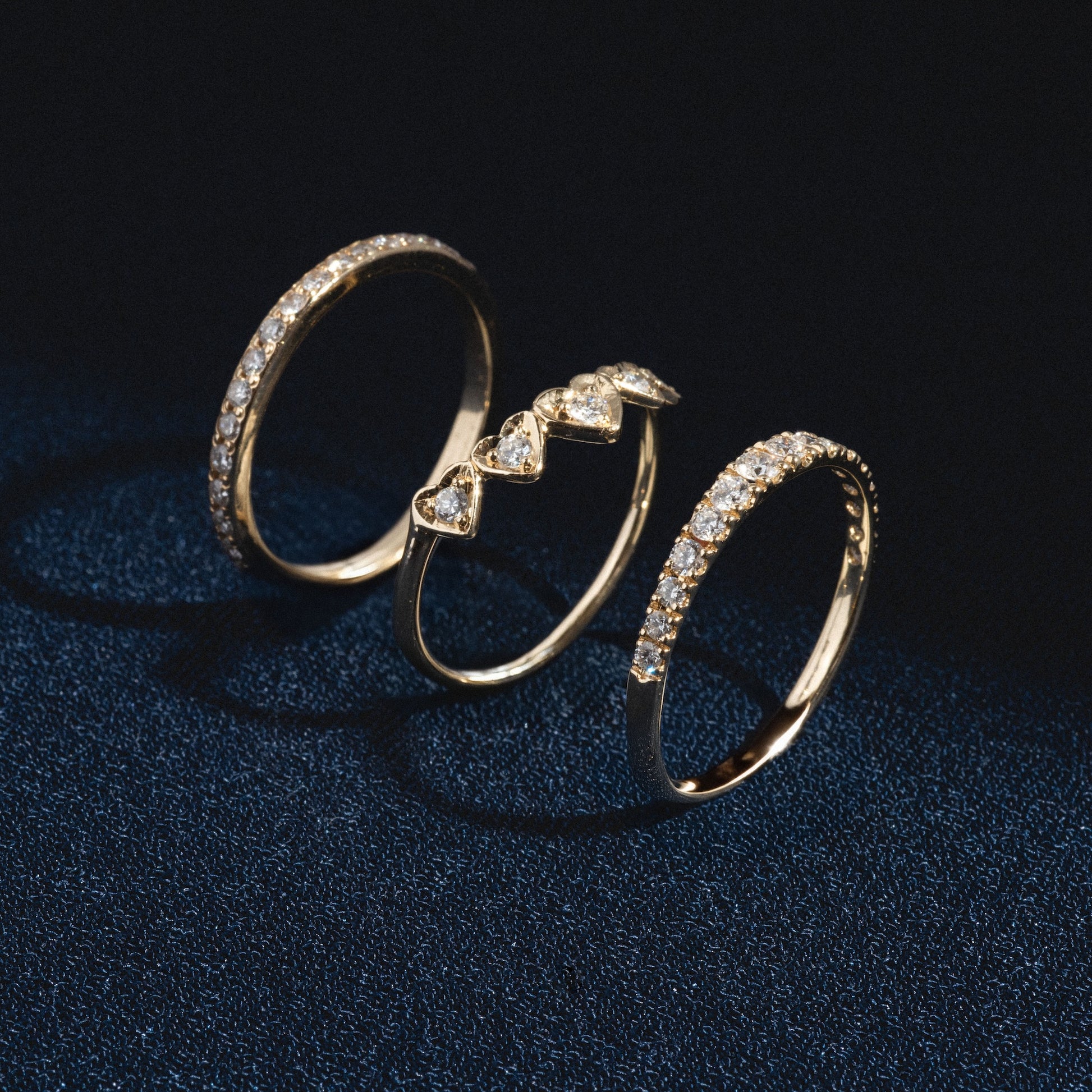The Radiant Hearts Band (outward band), a gold ring with diamond accents, is arranged on a dark textured surface, with light casting subtle shadows and highlighting the sparkle of its stones.
