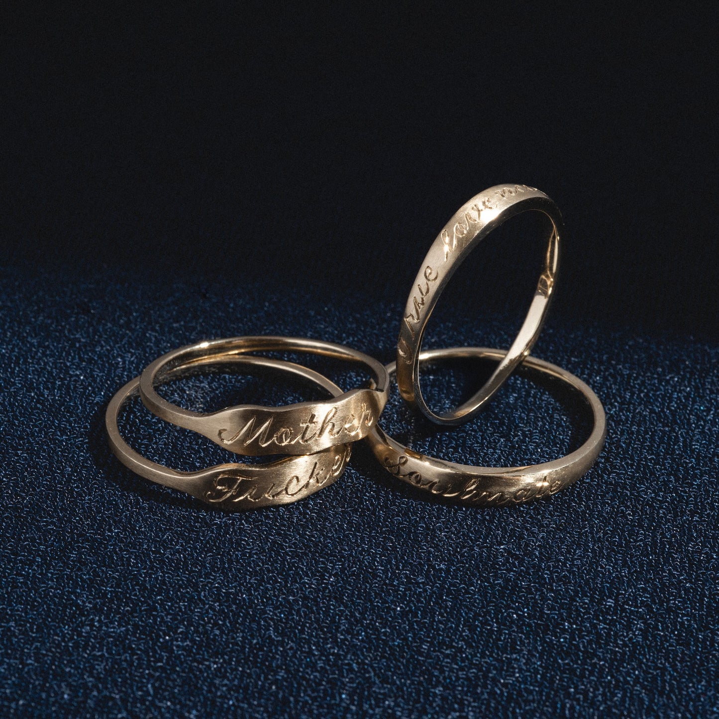 Four Soulmate Band gold rings with engraved words like Mother, Father, and partial inscriptions are displayed on a dark blue textured surface with dramatic lighting.