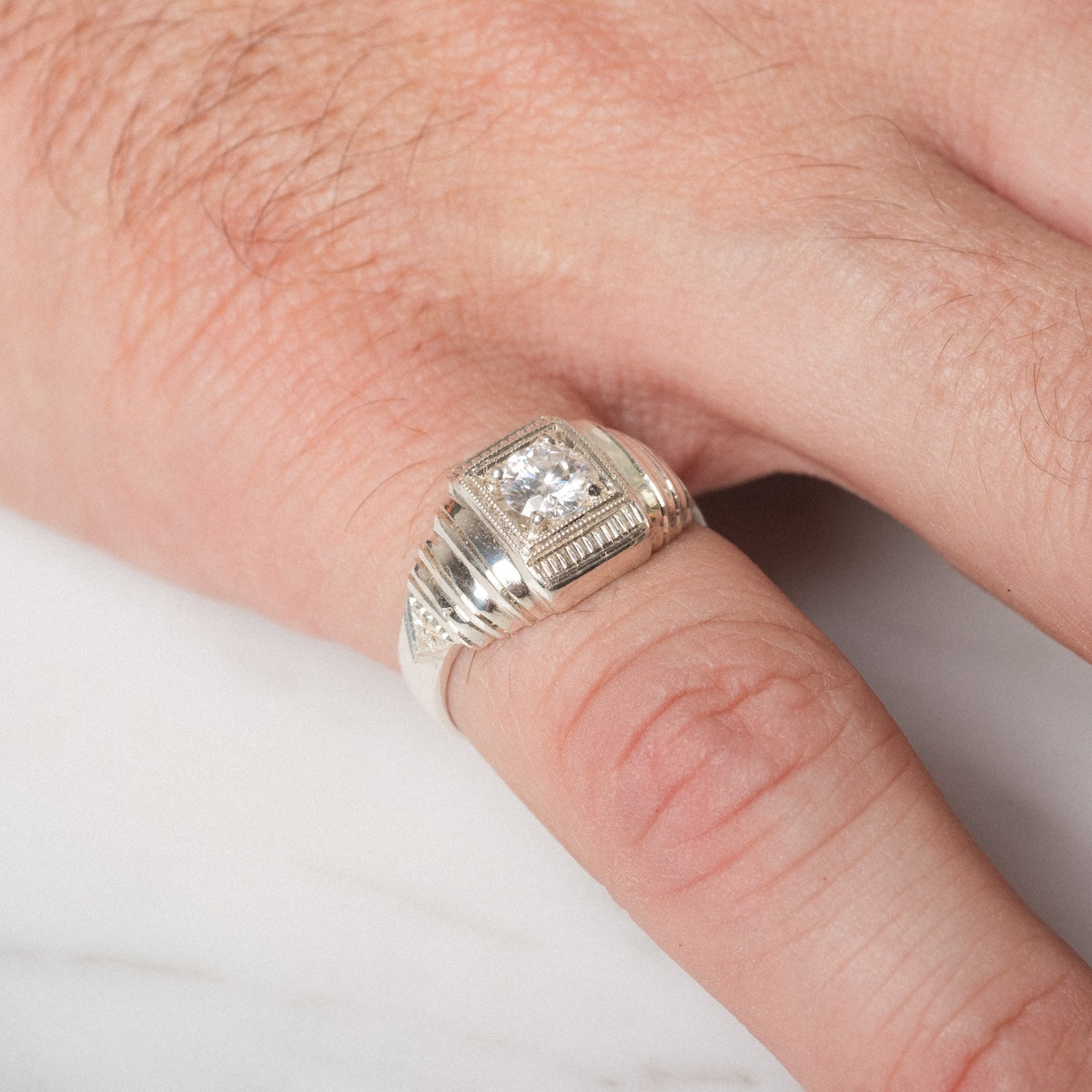 A close-up of a hand wearing the Sacred Ring, featuring a large square-cut clear gemstone set in a decorative silver ridged band, resting on a white surface.