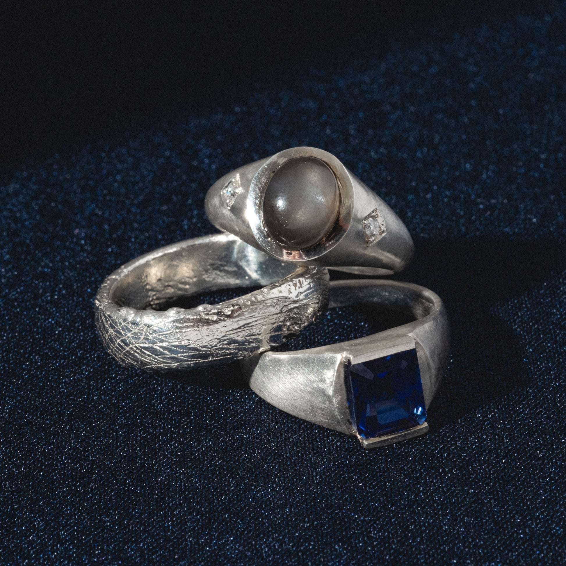 The Vailance Ring collection: three silver rings rest on dark blue fabric—one with a round gray stone, one with a textured band, and one showcasing a rectangular blue gemstone in a flat setting.