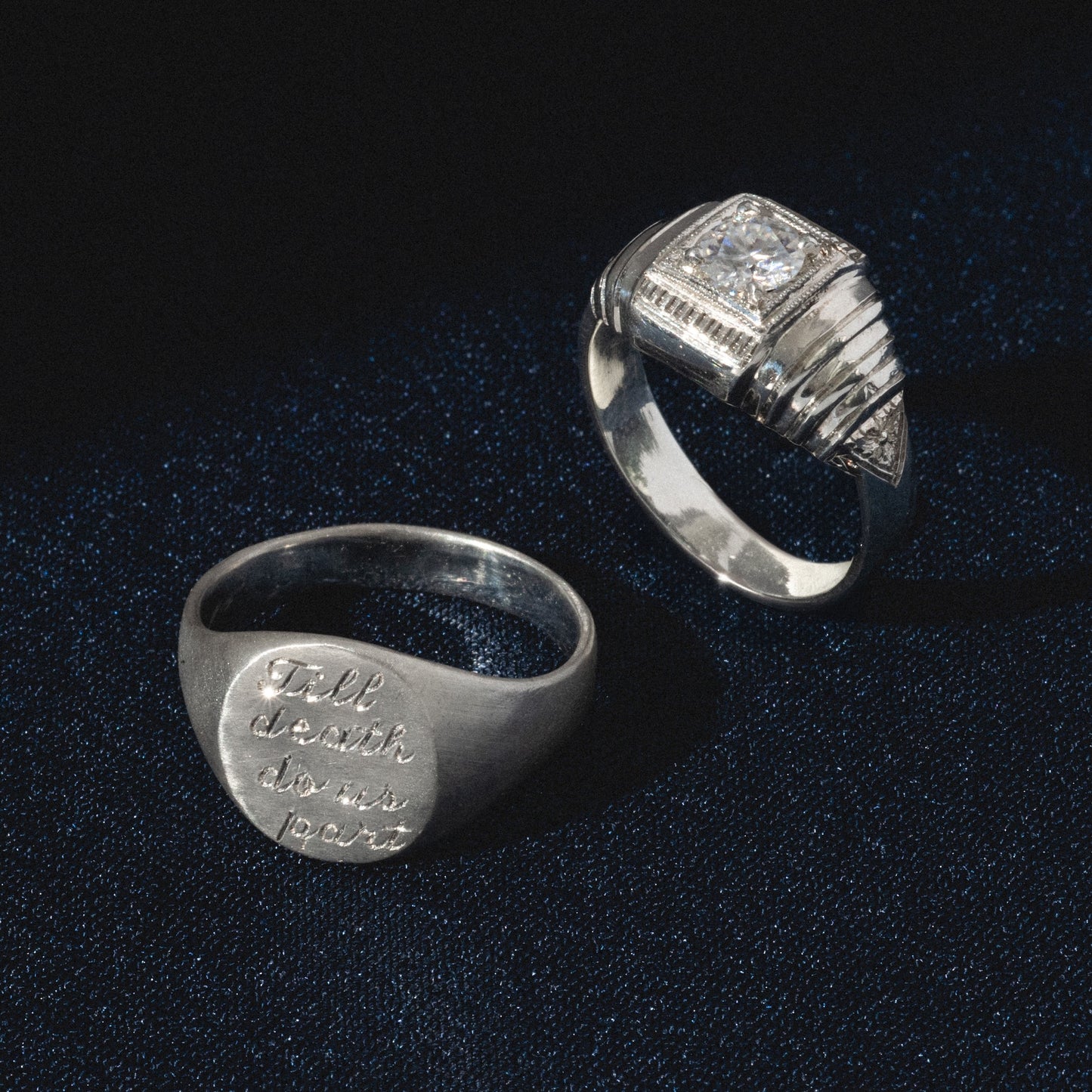 Two silver rings on dark fabric: one is adorned with a square-cut gemstone and geometric accents, while the other is the Till Death Do Us Part Signet Ring engraved with its signature phrase.