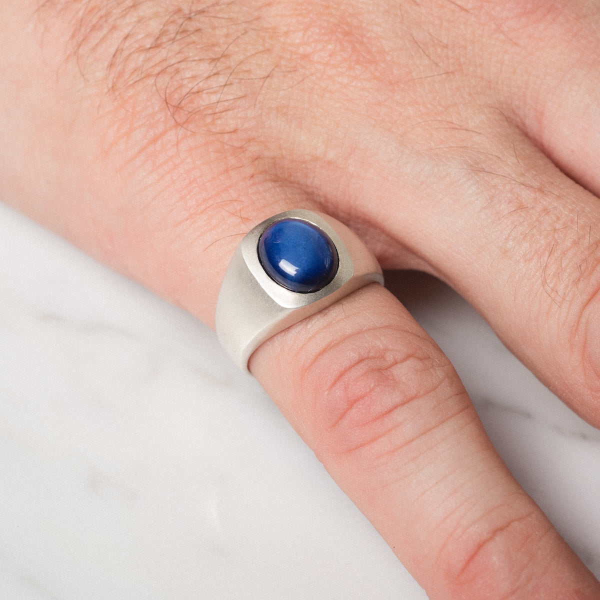 A hand wearing the Guardian Ring, featuring a large oval blue stone set in silver, rests on a white marble surface.