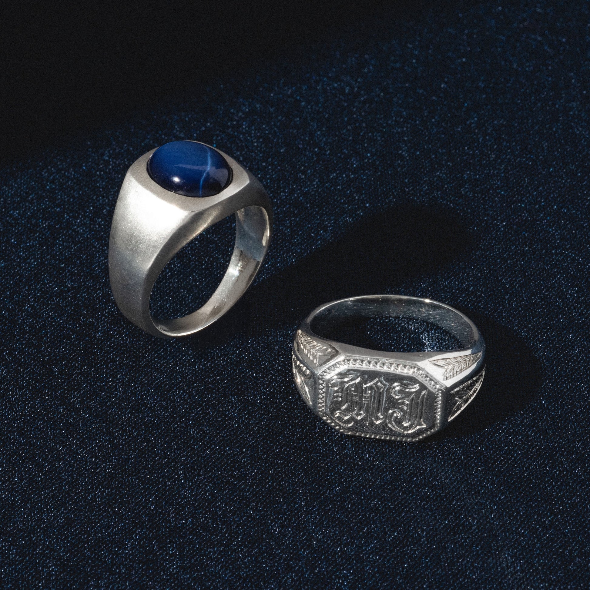 The Sovereign Signet Ring with a blue stone and an ornate engraved silver ring are displayed on dark blue fabric.