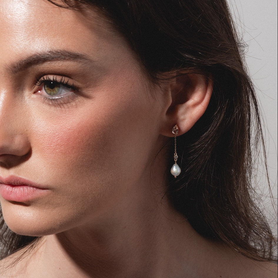 A woman with long brown hair and natural makeup looks to the left, wearing Solid Gold Citrine and Pearl Droplet Earrings that add a touch of elegance. Soft lighting highlights her clear skin and subtle blush.