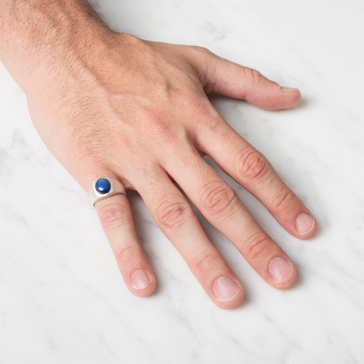 A hand rests on a white marble surface, wearing the Guardian Ring—a silver band featuring a round blue stone on the ring finger.