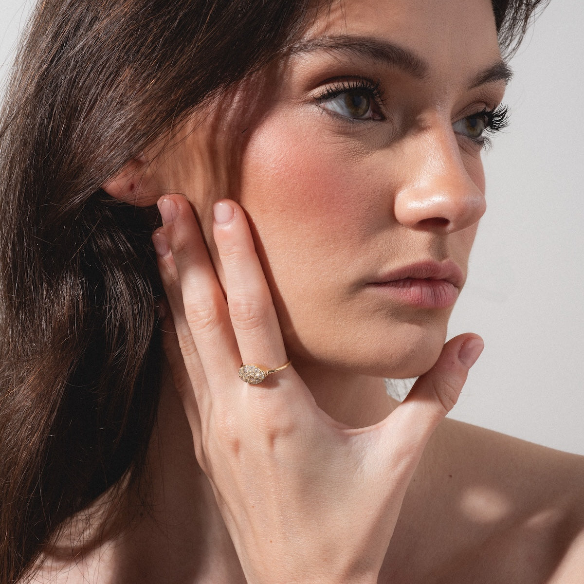 A woman with long brown hair, minimal makeup, and a thoughtful gaze touches her face while wearing the "Eternal Flame" Soul Ring, featuring a large, sparkling gemstone. She poses against a plain, light background.