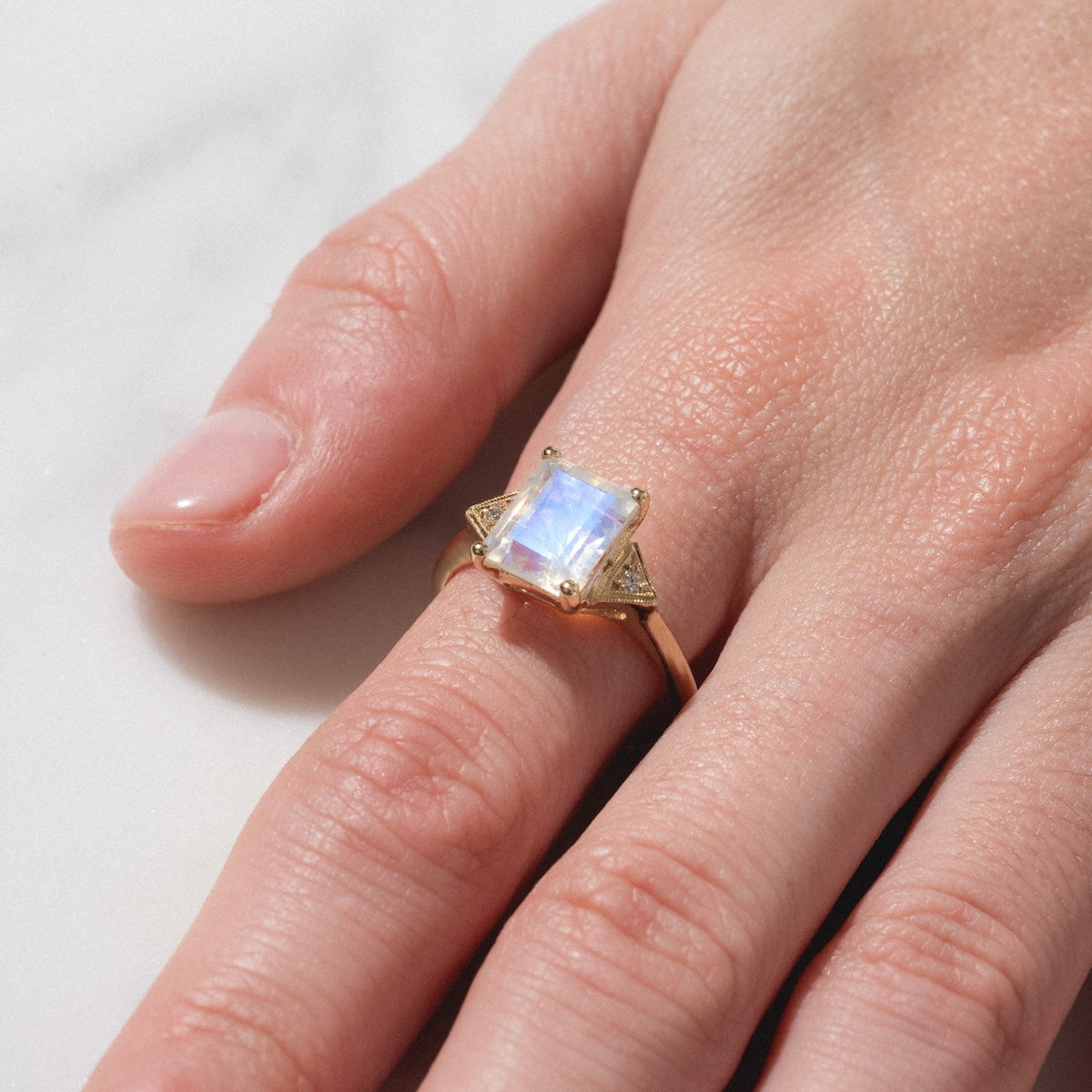 A hand adorned with the Celestia Ring, featuring a large square iridescent gemstone, rests on a light surface.