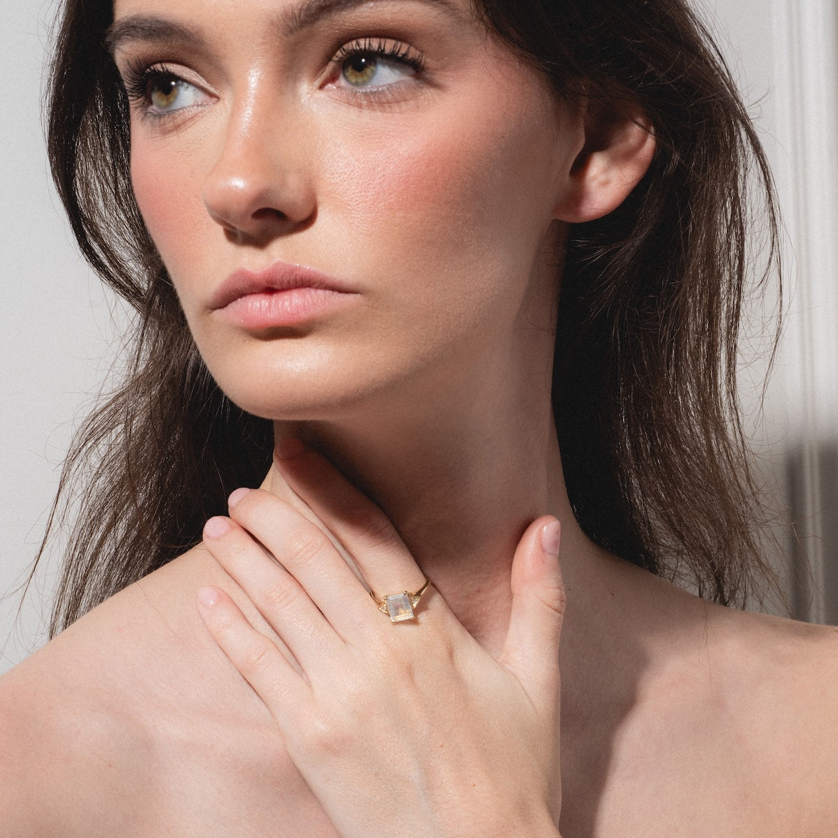 A woman with long brown hair touches her neck, showcasing the Celestia Ring—a bold, square-cut gemstone—on her finger. She wears natural makeup and poses against a neutral backdrop.