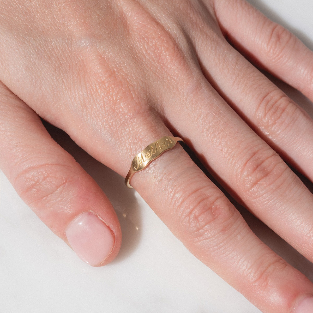 A close-up of a hand with neatly trimmed, unpolished nails rests on a white surface, showcasing the 14k Ripples Customizable ID Band with engraved text on the ring finger.