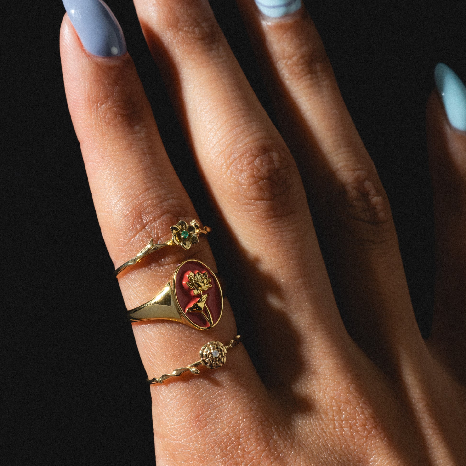 A close-up of a hand with long, light blue nails, wearing three gold rings—including a textured round ring and the Solid Gold Birth Flower Ring—set against a dark background.
