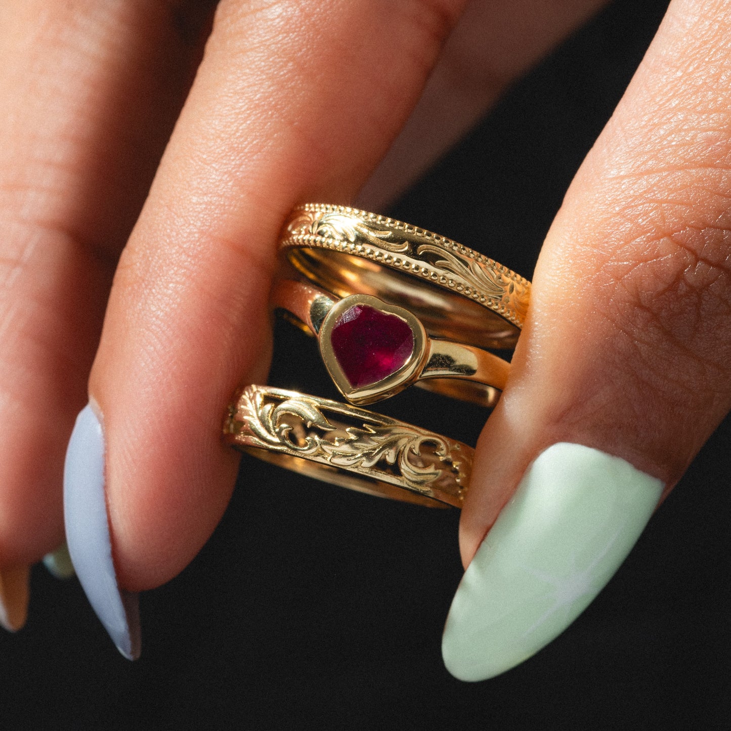 A hand with pastel green and blue nails displays three gold rings: the Solid Gold Bezel Set Ruby Heart Ring plus two engraved gold bands, making them perfect for everyday wear.