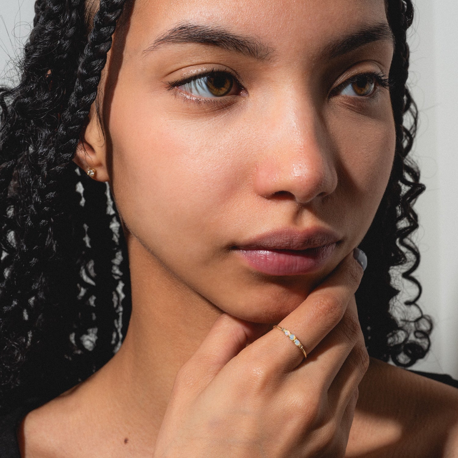 A young woman with curly hair gazes thoughtfully to the side, her chin resting on her hand. She wears the Solid Gold 2025 October Capsule Classic Opal Trio Statement Ring, radiating calm and contemplation.