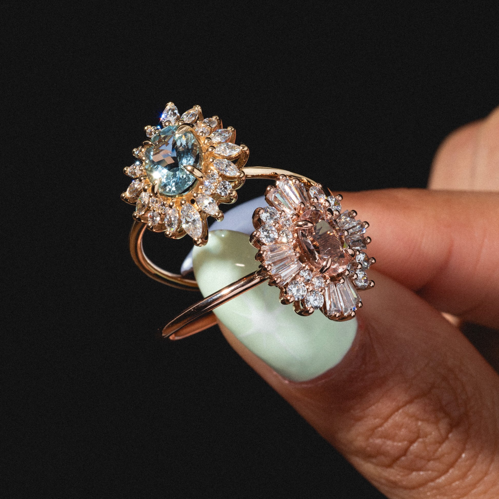 A hand with mint green nail polish holds two Lana Rings with Aquamarine & CZ, each in a 14k solid gold floral design featuring a central aquamarine gemstone and sparkling cubic zirconia, set against a black background.