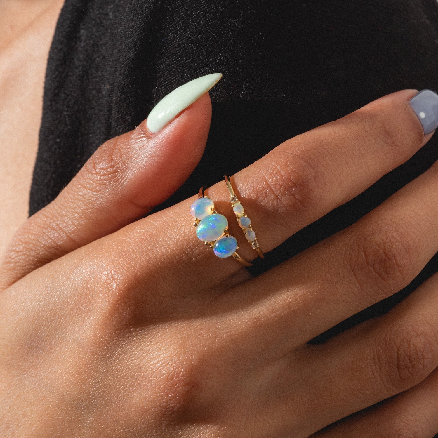 A hand with pale green and blue manicured nails showcases the Solid Gold 2025 October Capsule Opal Ring Set, featuring multiple iridescent opals, worn against a black fabric background. Comes with a free gift.
