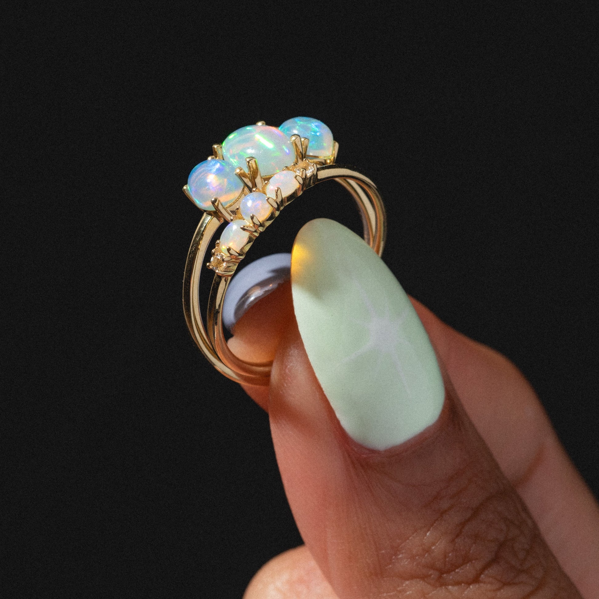 A hand with pastel green nails holds the Solid Gold 2025 October Capsule Opal Ring Set, featuring five iridescent opals, against a black background; includes a free gift.