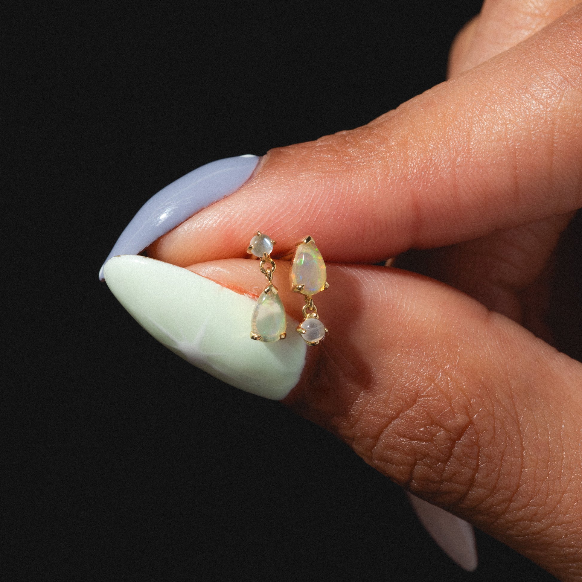 A hand with long, pastel nails holds the Solid Gold 2025 October Capsule Delicate Opal Drop Earrings—each features two petite teardrop-shaped opals, set in gold and showcased against a black background.