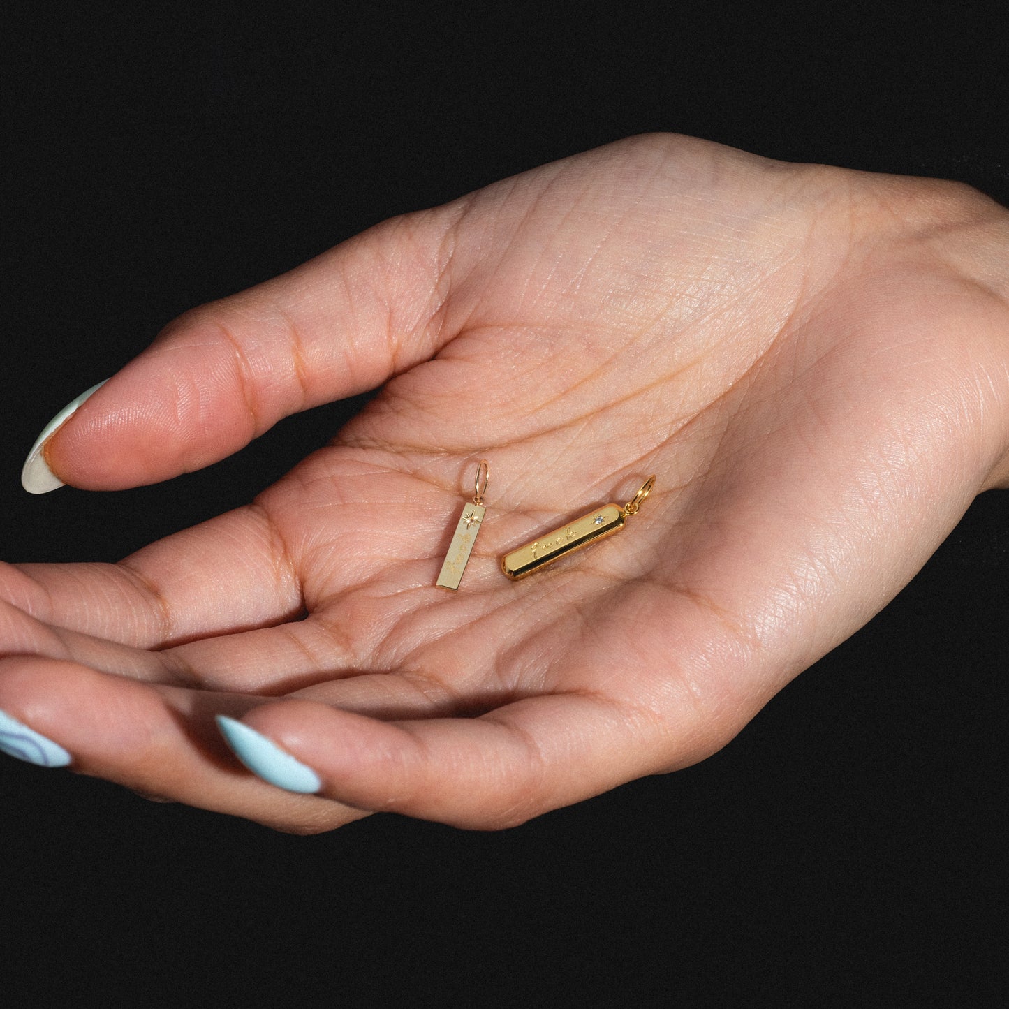 A close-up of a light-skinned hand with long, light blue nails holding two Tiny Fuck Charm gold pendants, set against a black background.