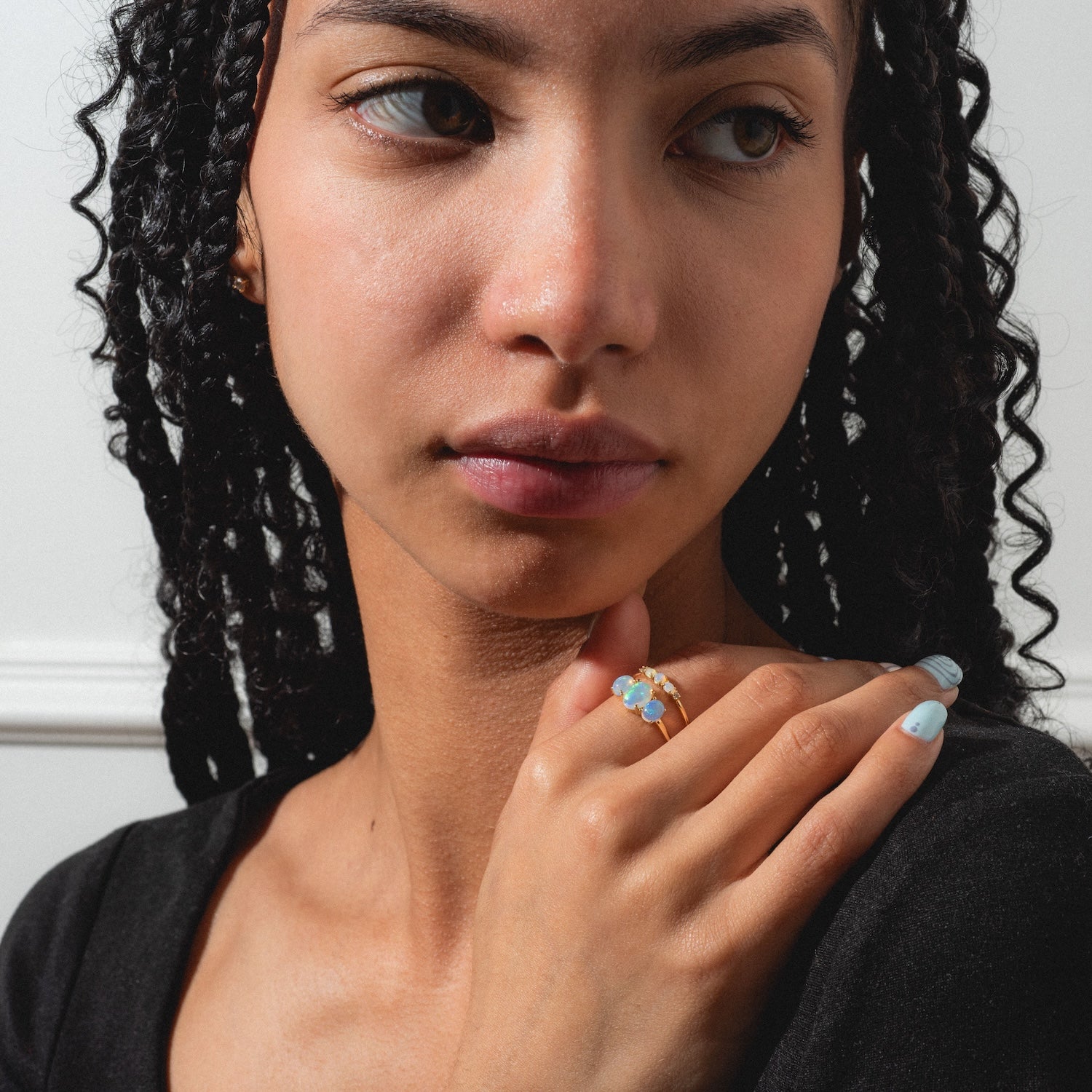 A woman with braided hair and light blue nails looks to the side, hand on shoulder. She wears the Solid Gold 2025 October Capsule Opal Ring Set with Free Gift and a black top, set against a light background.