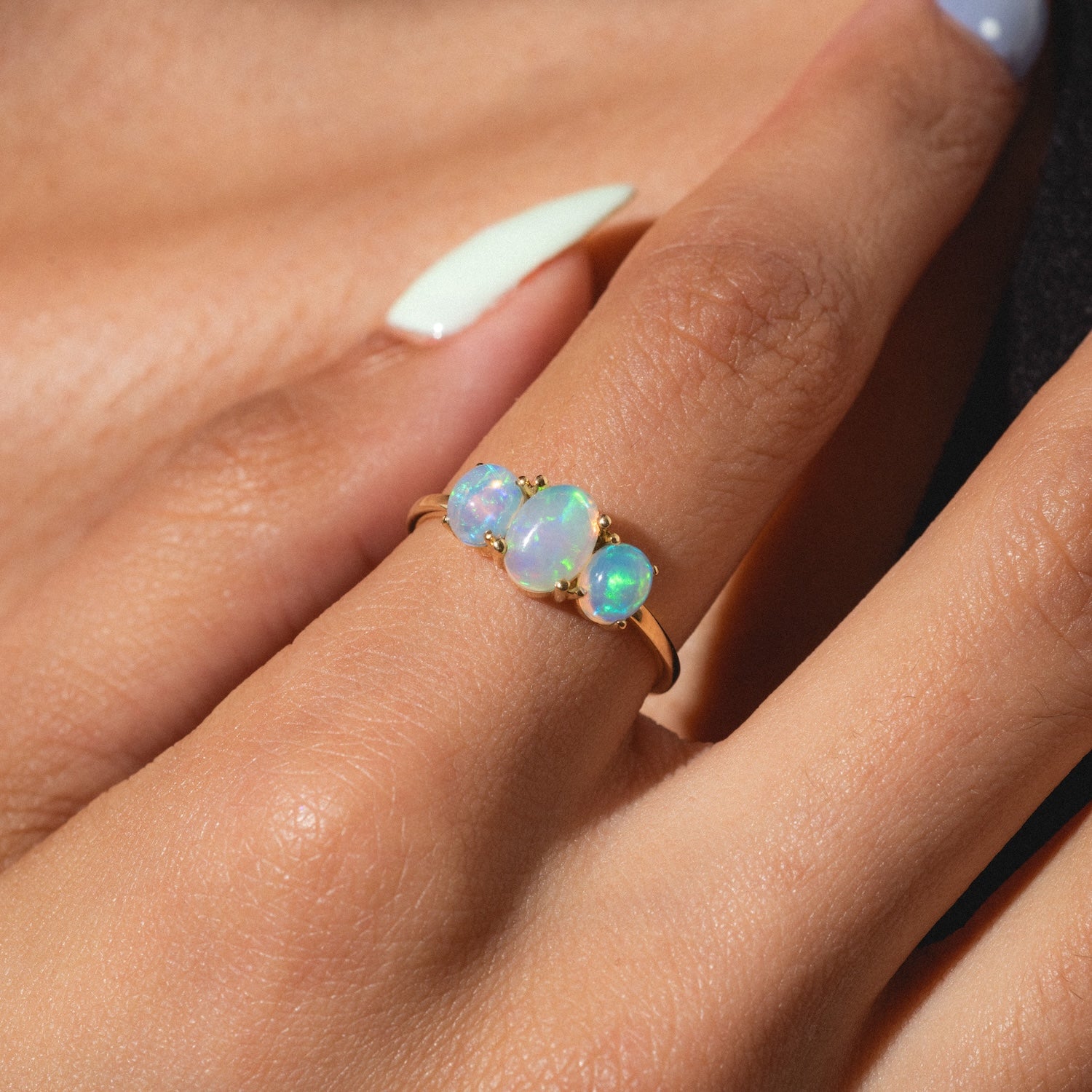 A hand with manicured nails wears the Solid Gold 2025 October Capsule Opal Statement Set—three oval opal stones with iridescent colors. The softly blurred background highlights the free gift included.