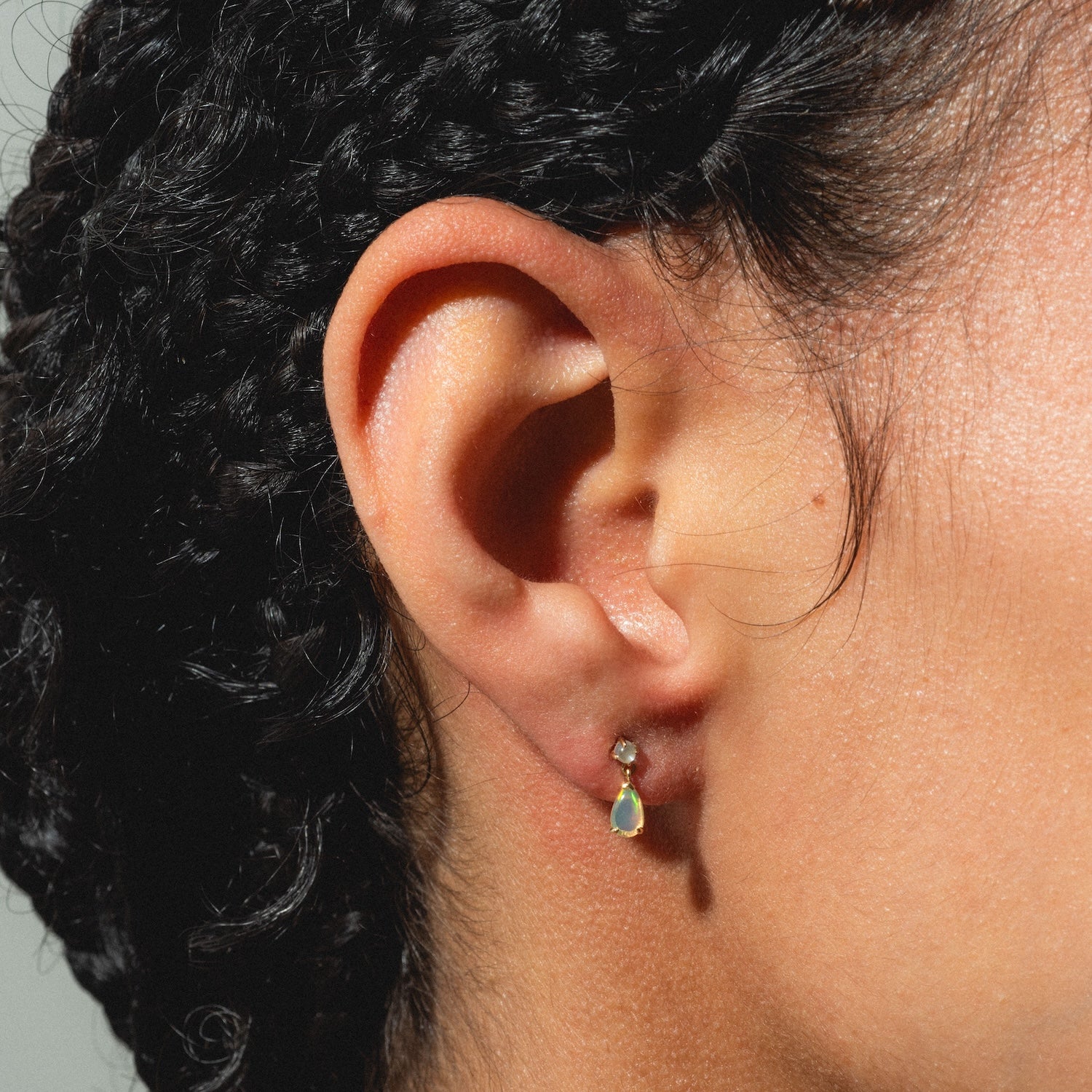 Close-up of a person's ear with short, curly hair, wearing an earring from the Solid Gold 2025 October Capsule Opal Statement Set: a clear stone above a dangling teardrop-shaped green gem. Includes free gift.