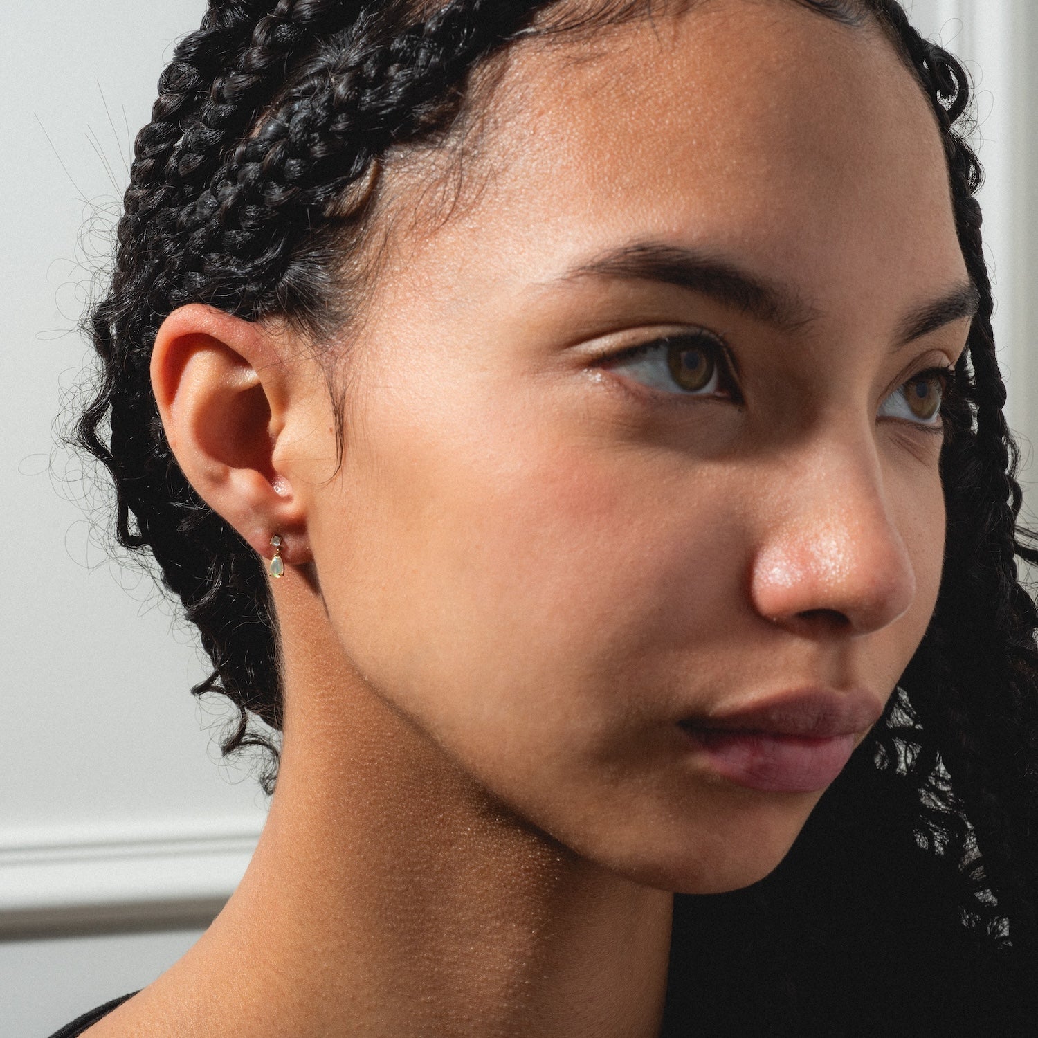A young woman with braided hair gazes thoughtfully to the side, wearing Solid Gold 2025 October Capsule Delicate Opal Drop Earrings against a light background.