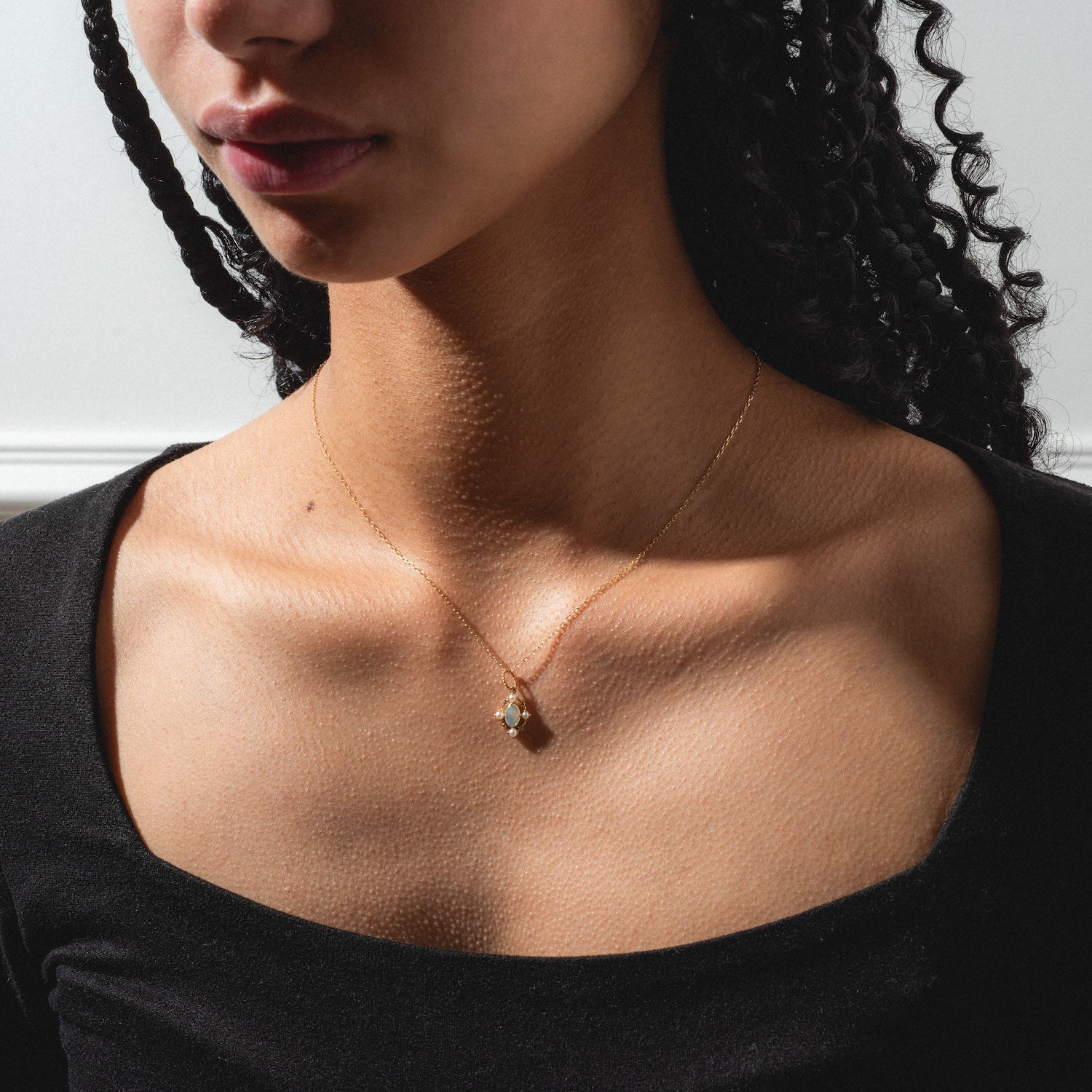 A woman with curly hair wears a black top and the Solid Gold 2025 October Capsule Vintage Inspired Opal Pendant Necklace, its delicate pendant glimmering in soft light that highlights her collarbone and textured skin.