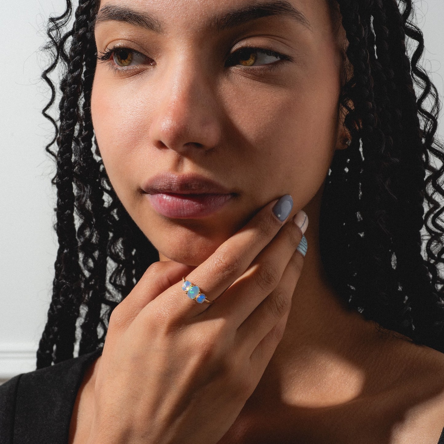 A woman with long braided hair gazes thoughtfully to the side, resting her chin on her hand as she wears the Solid Gold 2025 October Capsule Classic Opal Trio Statement Ring, its opals glowing softly in the natural sunlight.