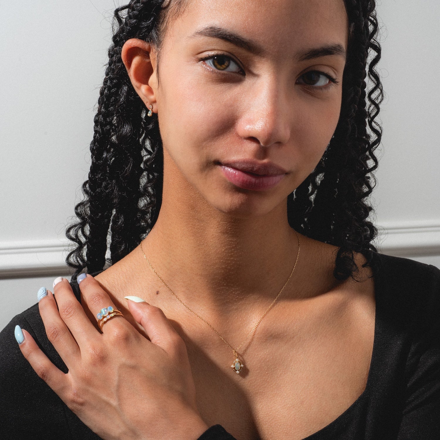 A woman with curly black hair wears the Solid Gold 2025 October Capsule Opal Full Set—pendant necklace, three-stone ring, and stud earrings. She has light blue nail polish and poses against a white wall.