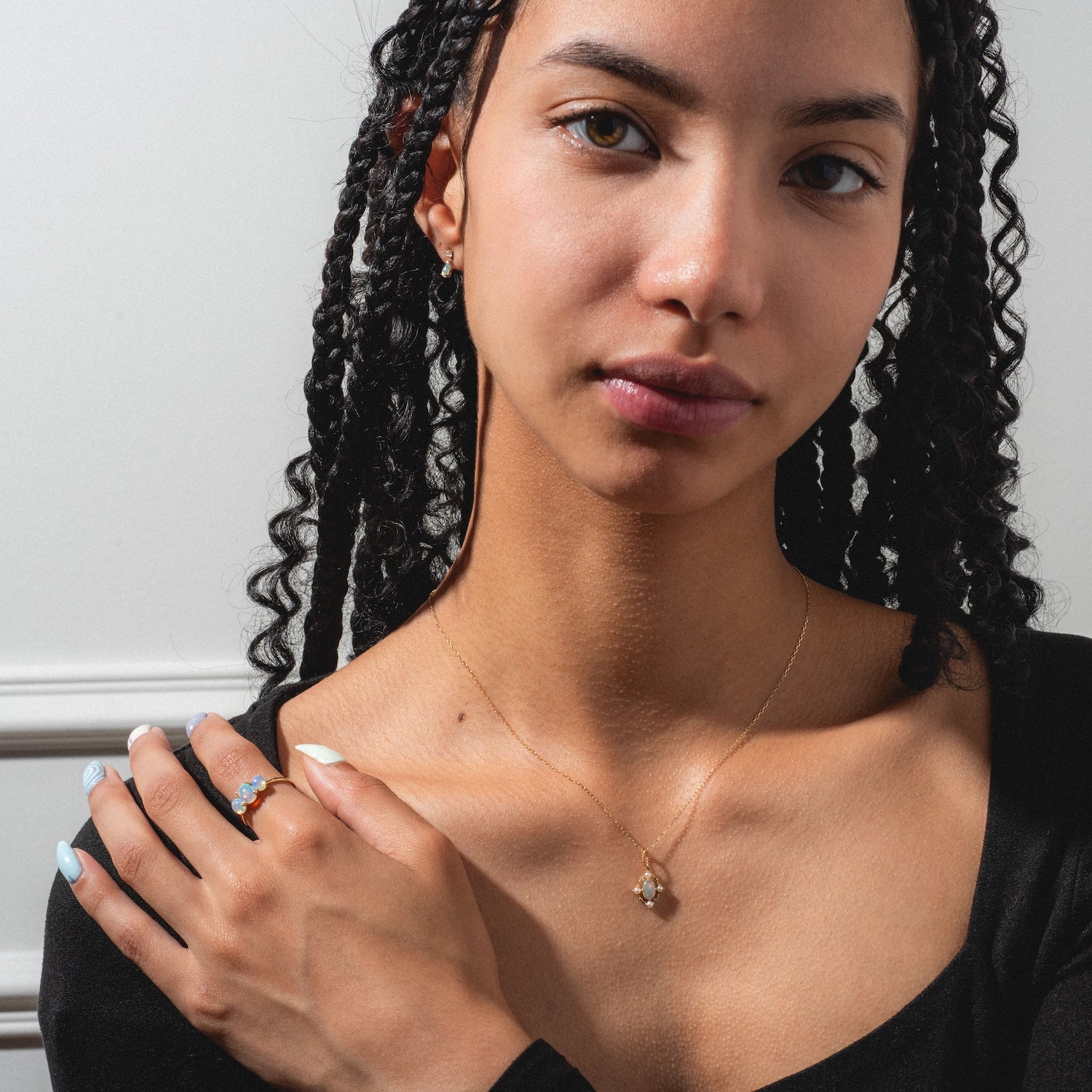 A woman with braided hair wears the Solid Gold 2025 October Capsule Opal Statement Set and a gold ring with blue stones. Sporting natural makeup, light blue nail polish, and a black top, she stands against a white wall. Free gift included.