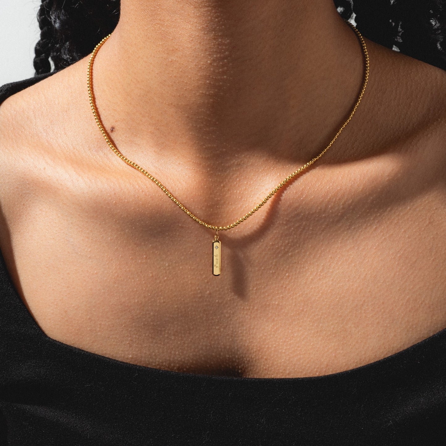 A person wears a gold "Tiny Fuck Charm" rectangular pendant necklace with a black off-the-shoulder top. The photo highlights their neck and collarbone area.